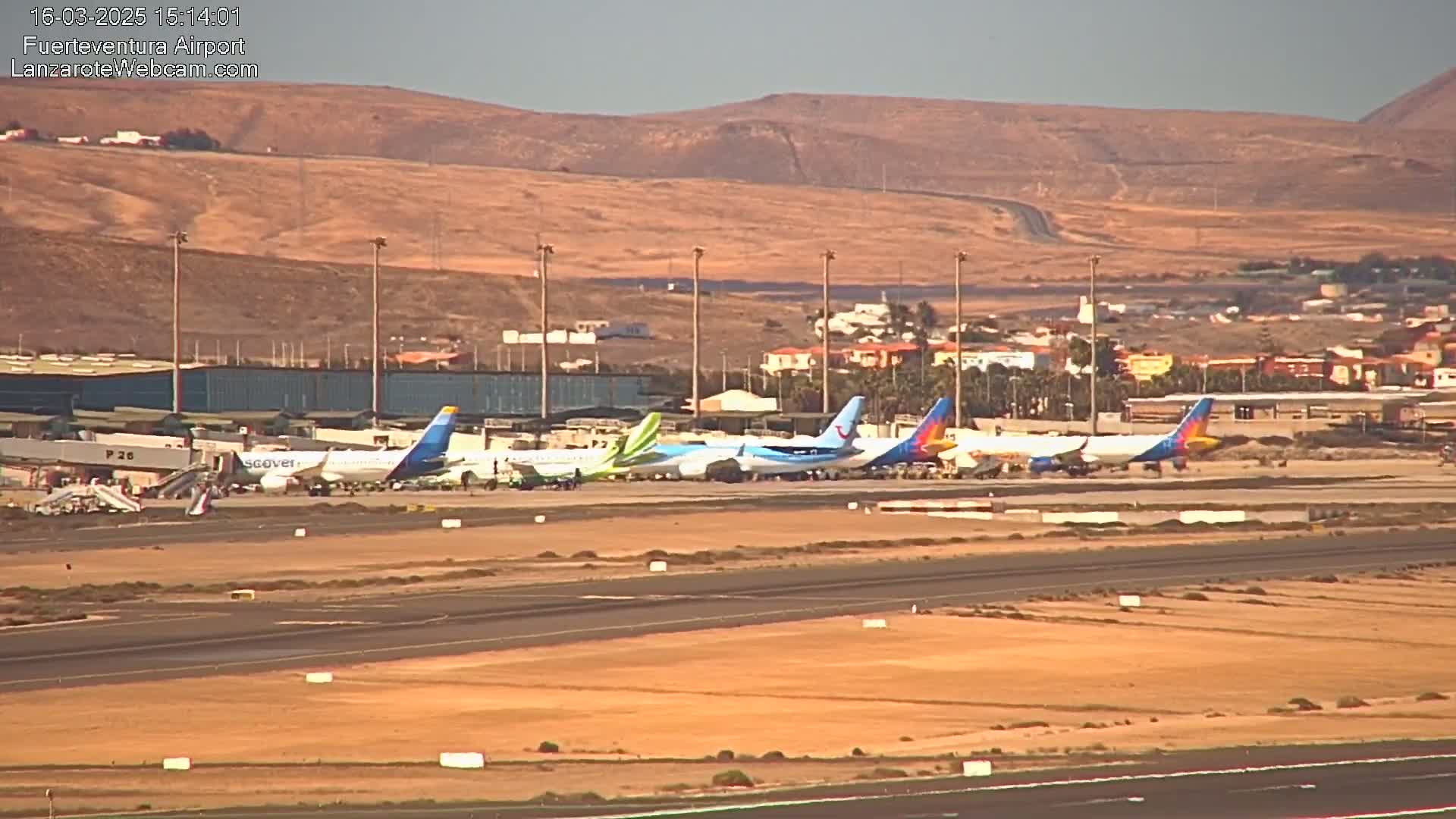 Several airplanes are parked on a tarmac at an airport under a clear, sunny sky, with a brown, hilly landscape visible in the background.