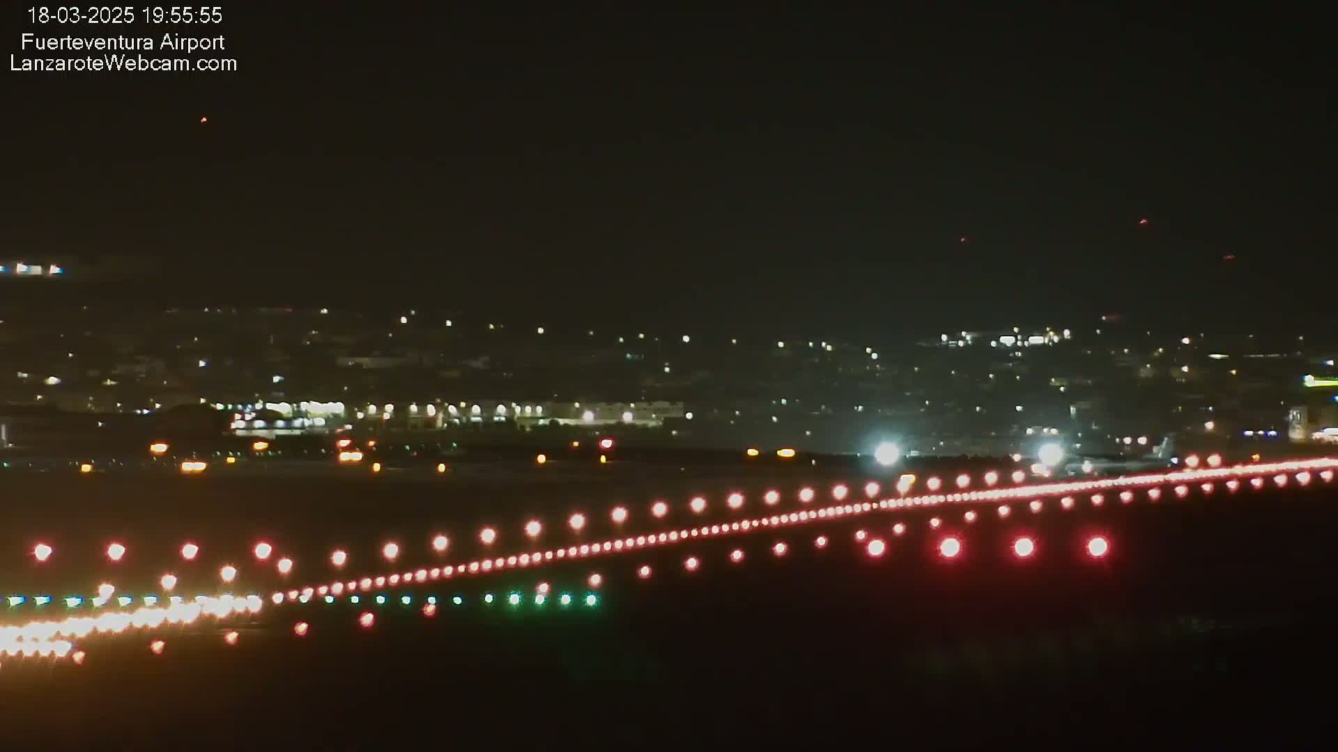 A nighttime view of an airport runway with its lights illuminated, and a city skyline faintly visible in the distance under a clear, dark sky.