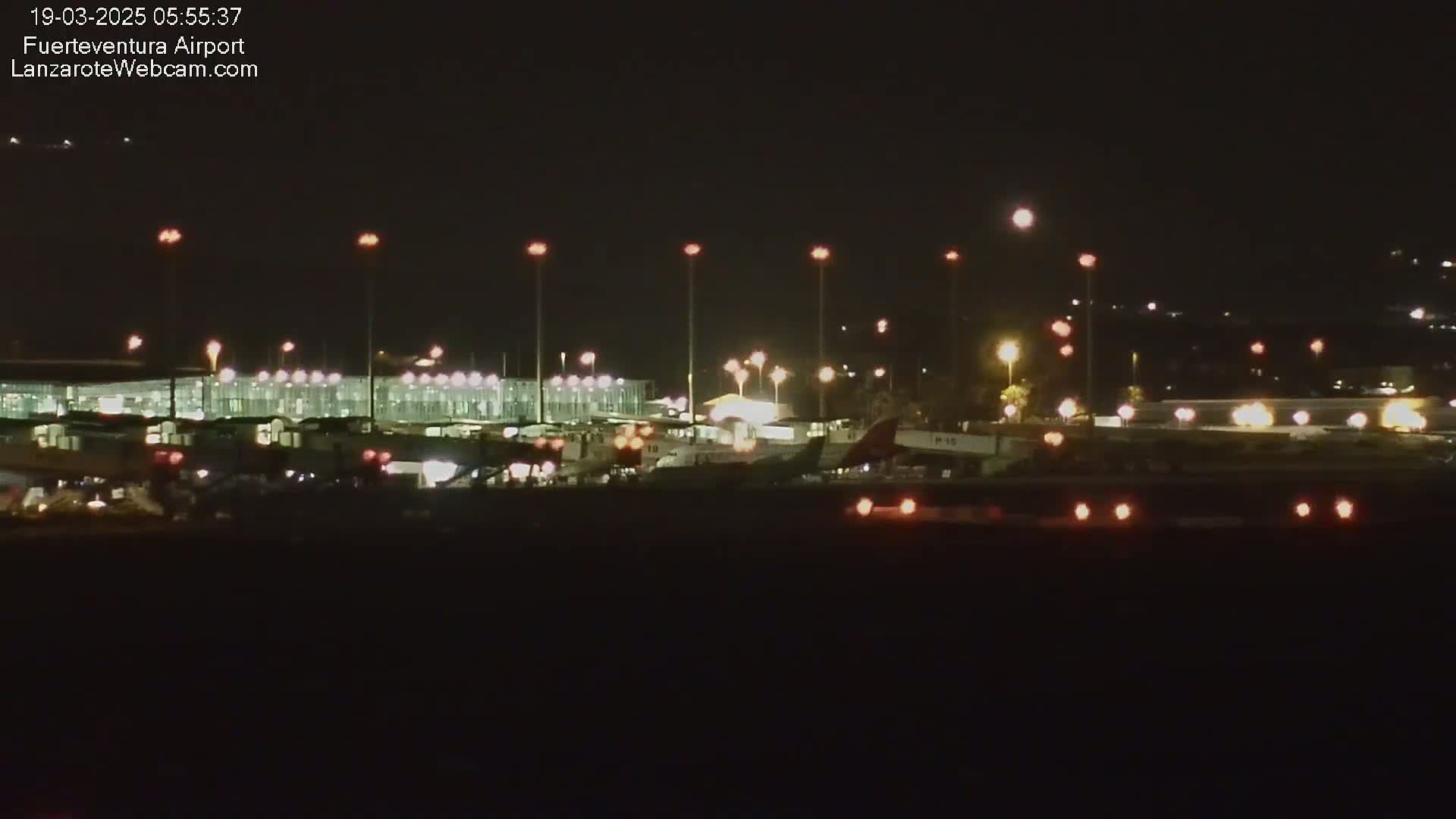 A nighttime view of an airport tarmac with several airplanes parked near a brightly lit terminal building under a clear, dark sky.