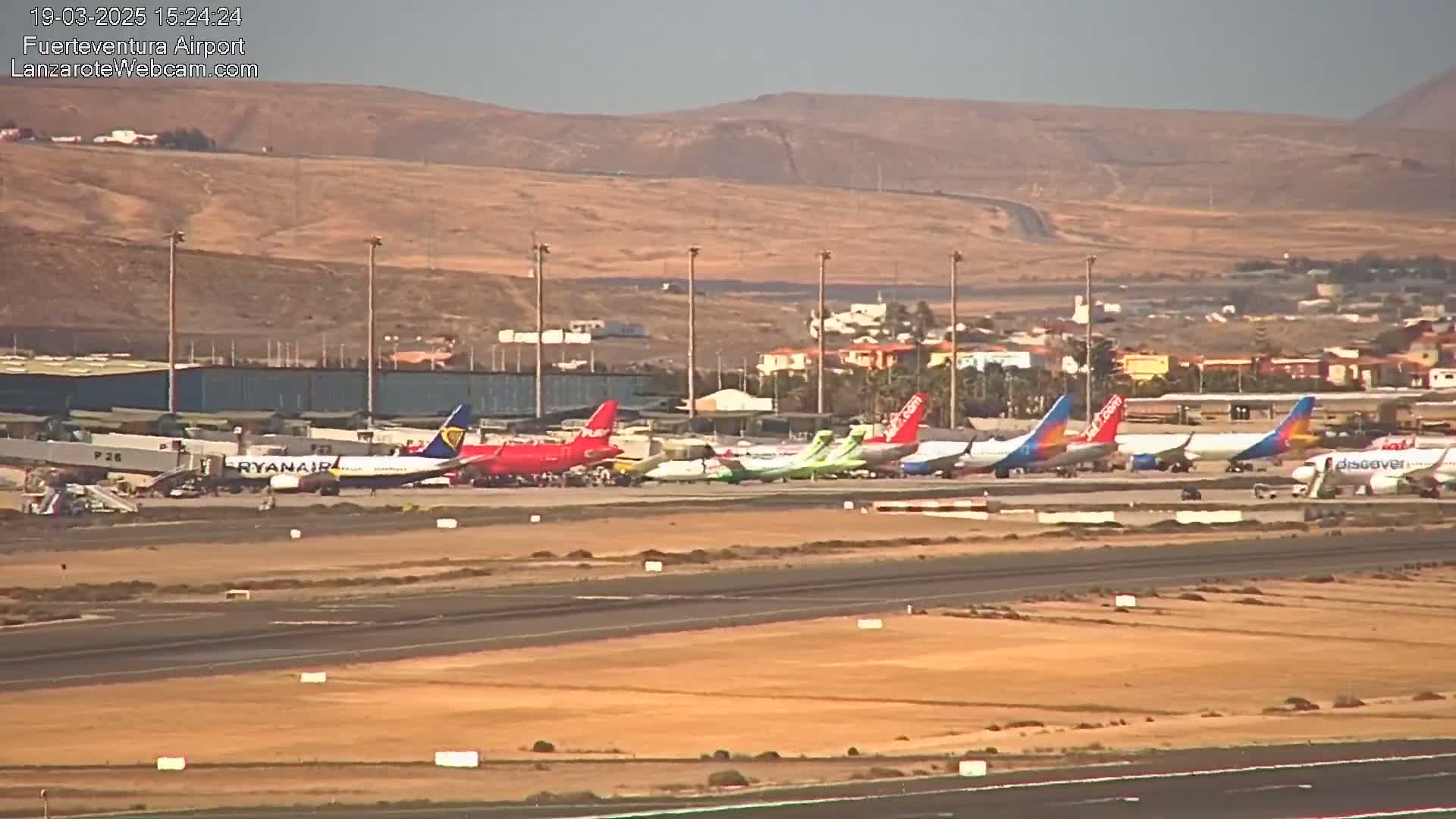 Several passenger planes are parked at an airport tarmac under a hazy sky, with arid hills visible in the background.