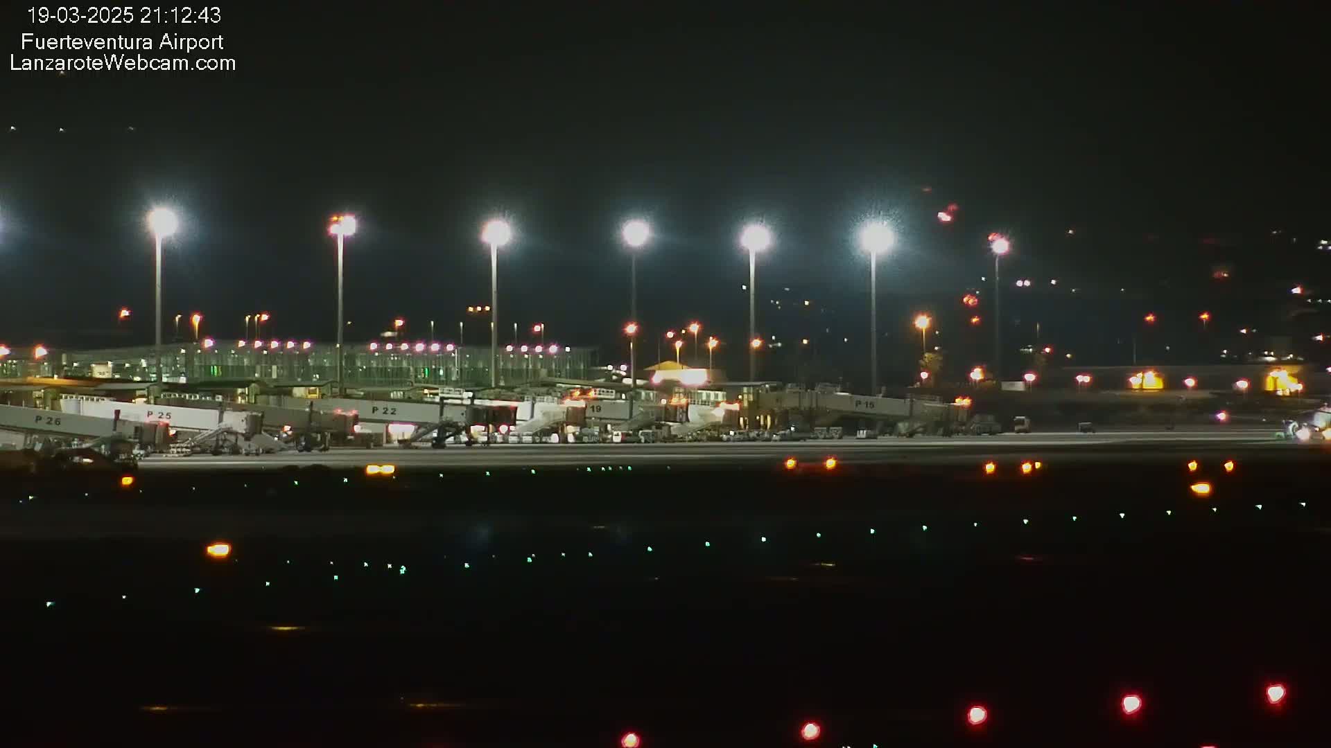A brightly lit airport tarmac at night shows several gates with passenger boarding bridges, ground support equipment, and runway lights.