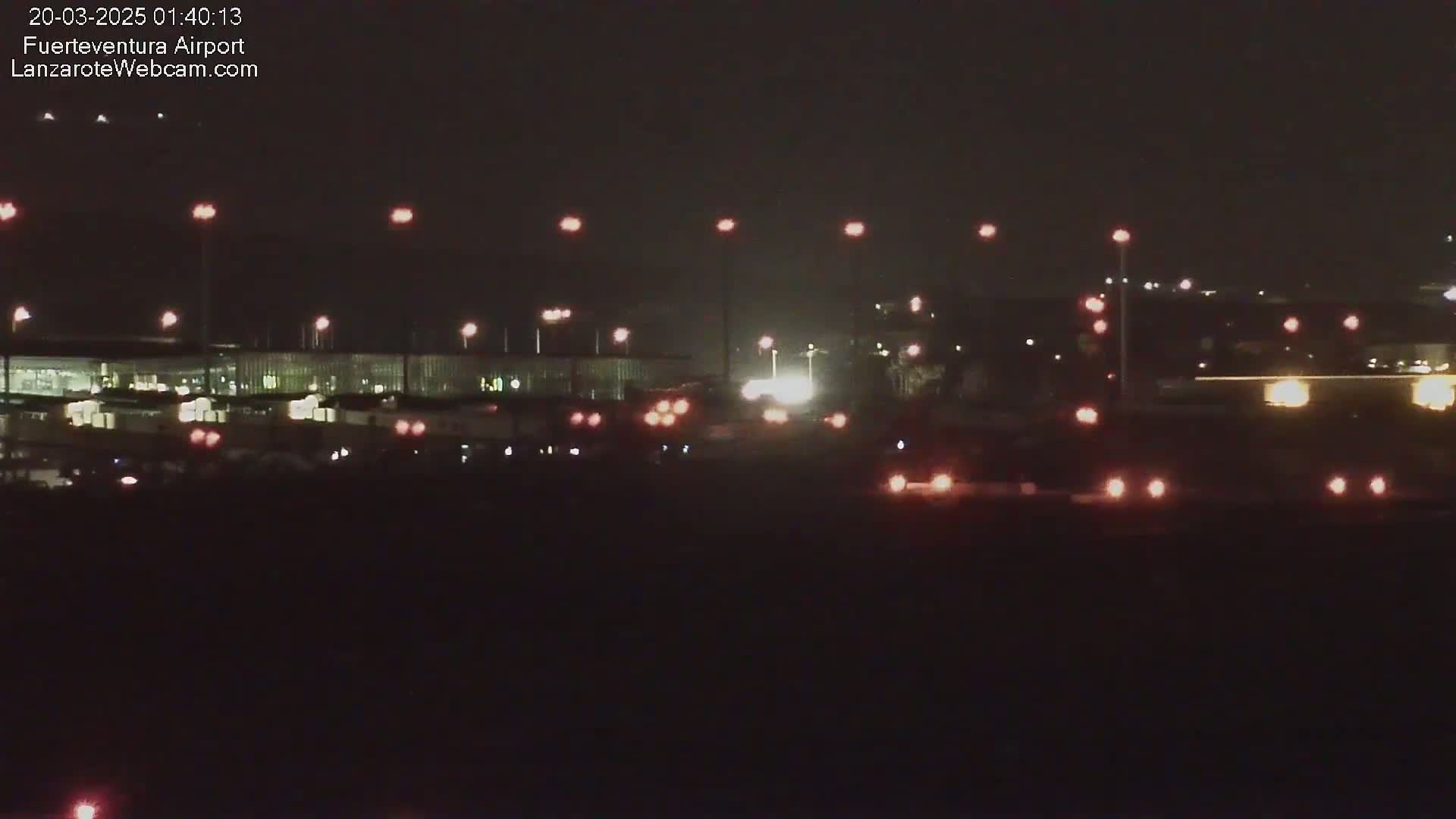 A nighttime long shot of an airport terminal building with numerous lights illuminating the tarmac and surrounding areas under a dark sky.