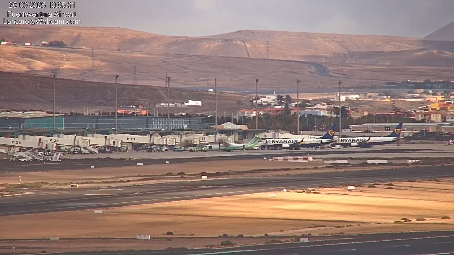 An airport tarmac with several planes, jet bridges, and airport buildings is visible under a partly cloudy sky, with arid hills in the background.