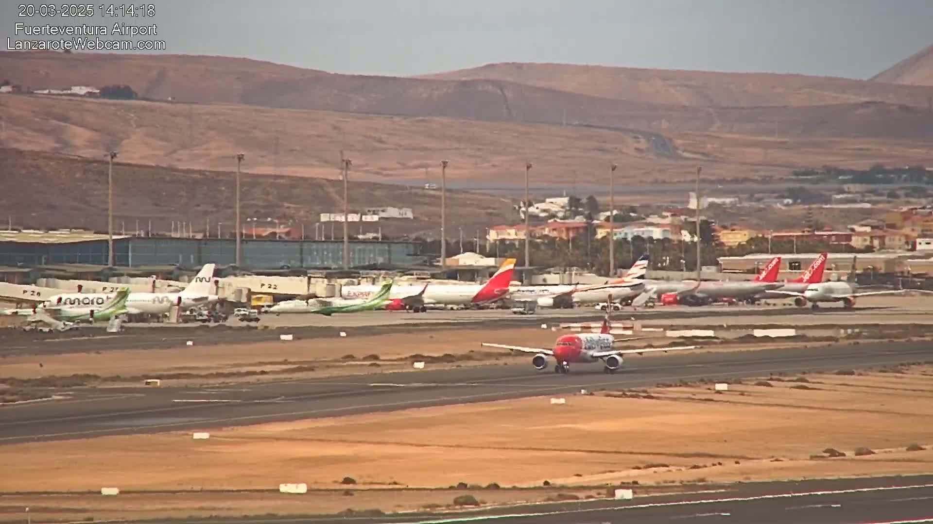 A red and white airplane taxis on a runway at an airport under a hazy sky, with numerous other airplanes parked nearby.