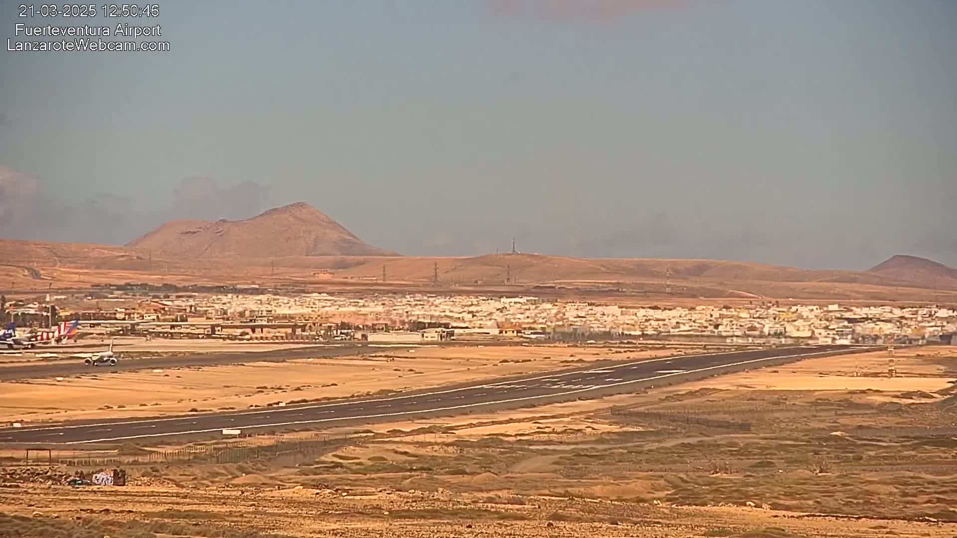 An airport runway stretches across a flat, arid landscape under a hazy sky, with a small town and distant hills visible.