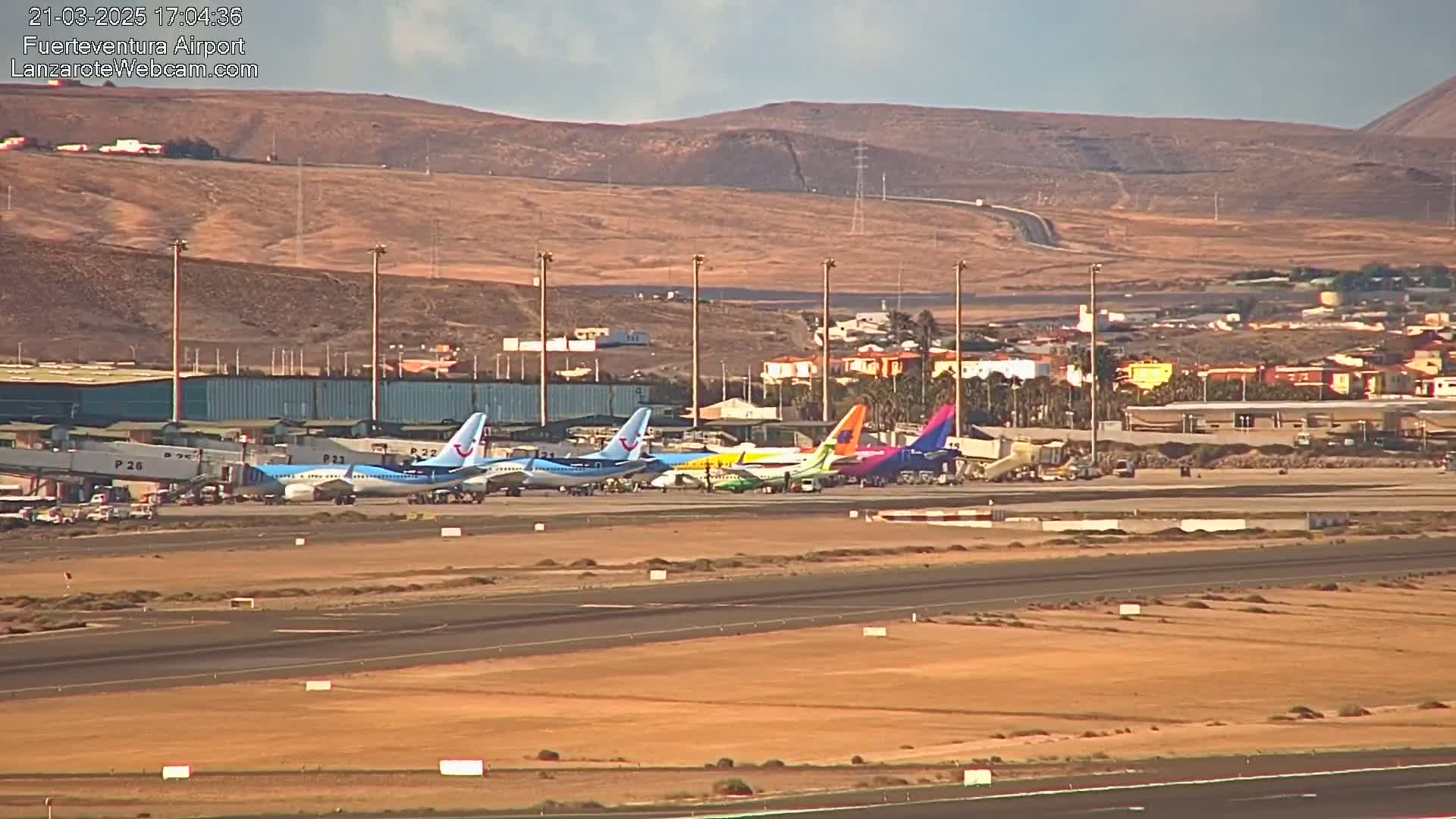 Several airplanes are parked at an airport tarmac next to a terminal building under a clear sky, with brown hills visible in the background.