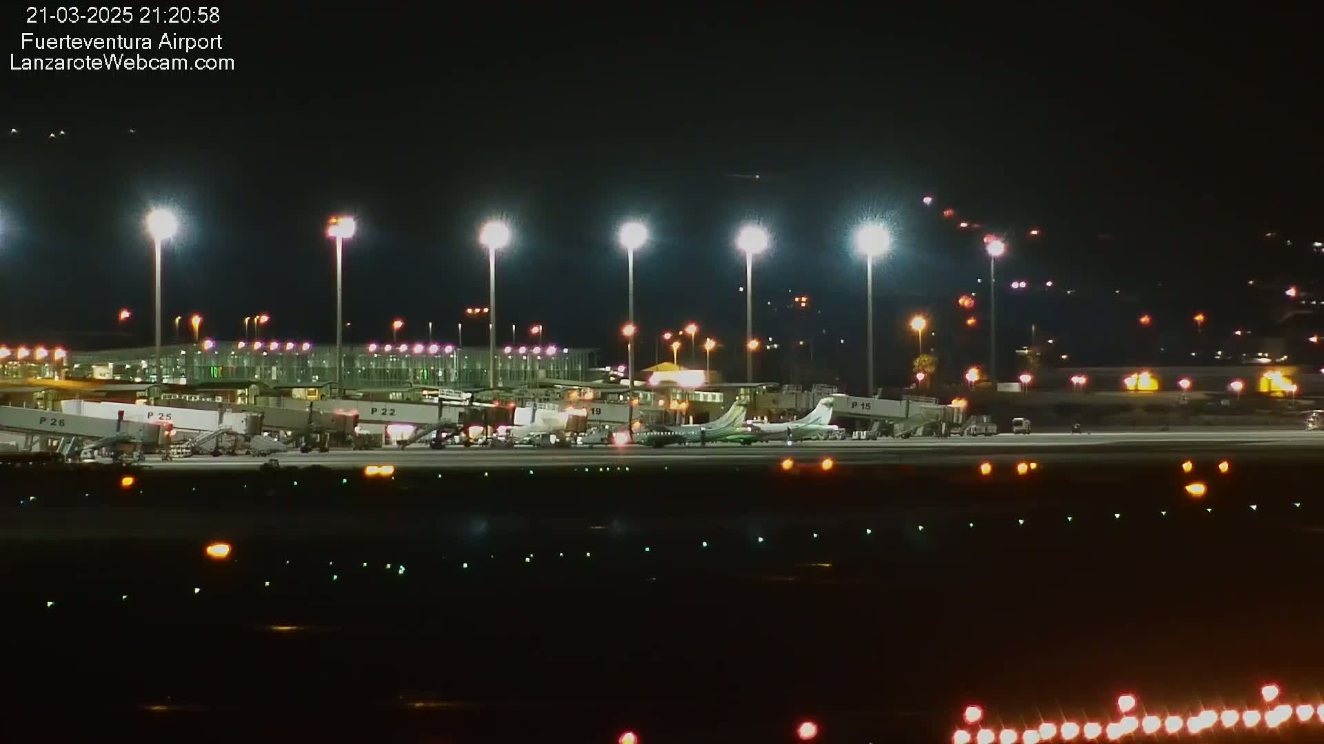 A brightly lit airport tarmac at night shows several airplanes parked at gates with jet bridges attached, under a clear, dark sky.