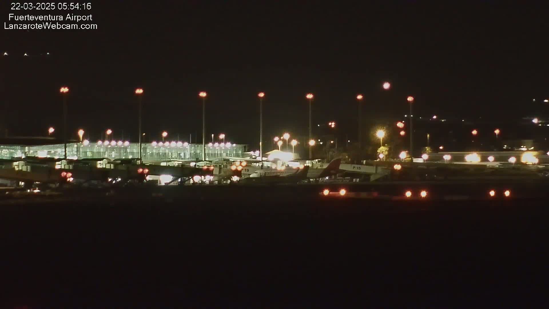 A brightly lit airport at night shows several airplanes parked near a terminal building under a clear, dark sky.
