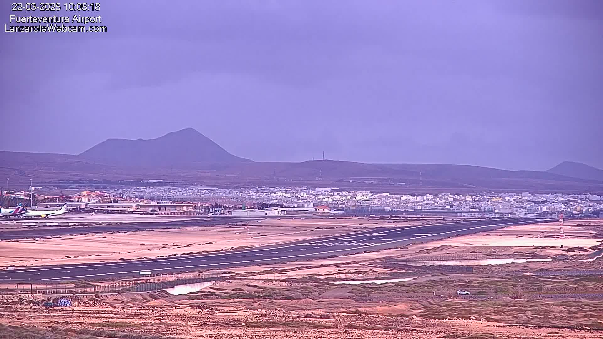 An airport runway and surrounding buildings are visible under an overcast sky, with volcanic hills in the background.