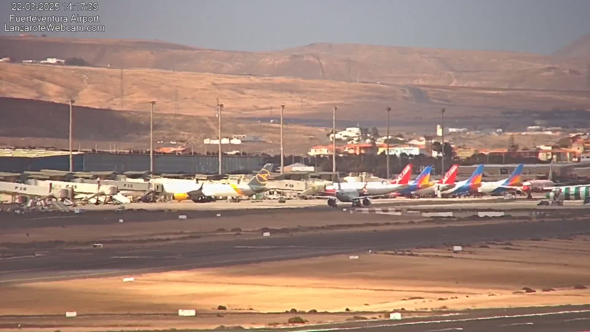 Several airplanes are parked on an airport tarmac, with a hazy sky and tan-colored hills in the background.
