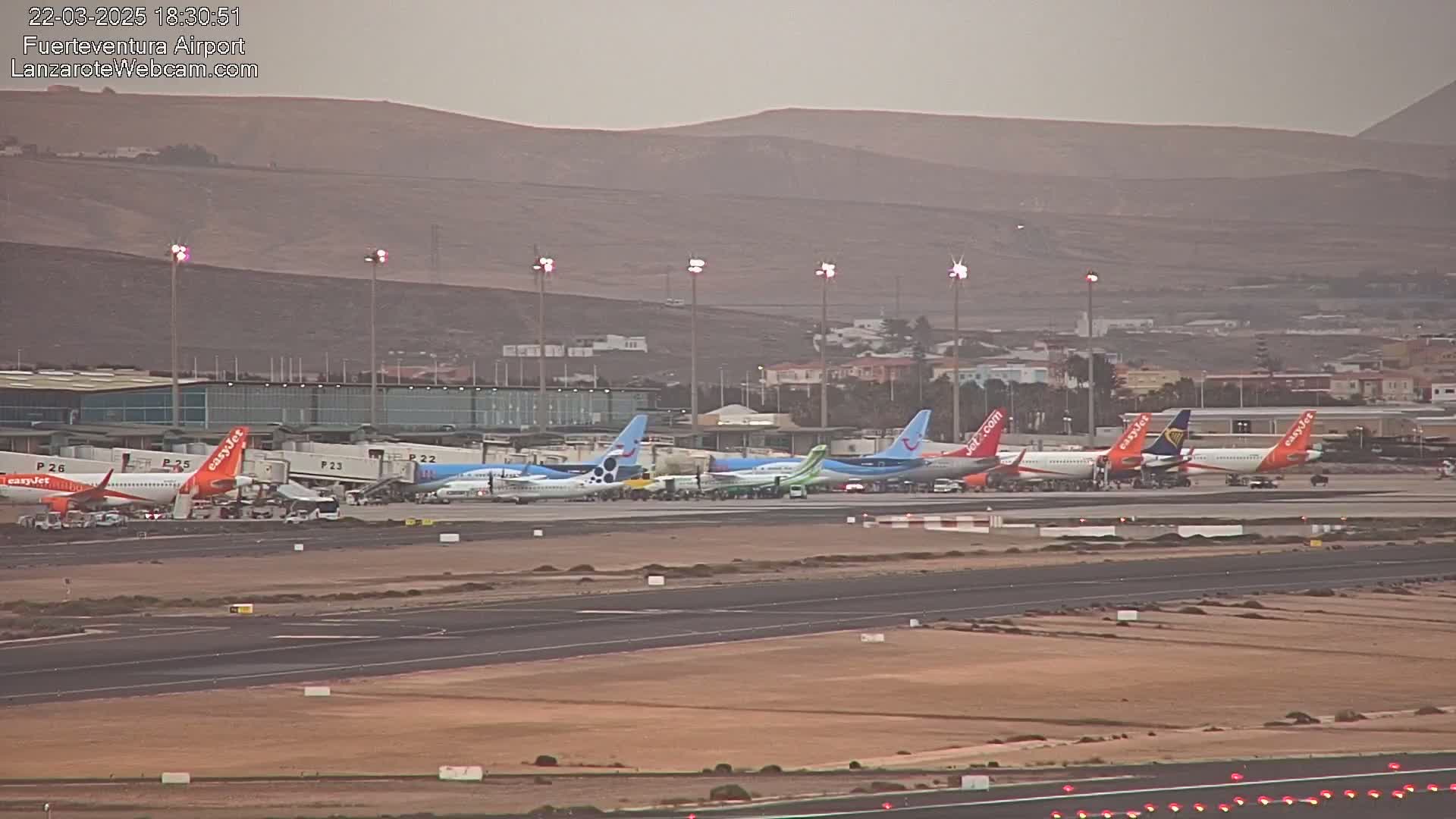 Several airplanes are parked at an airport terminal under hazy, overcast skies, with a range of hills visible in the background.