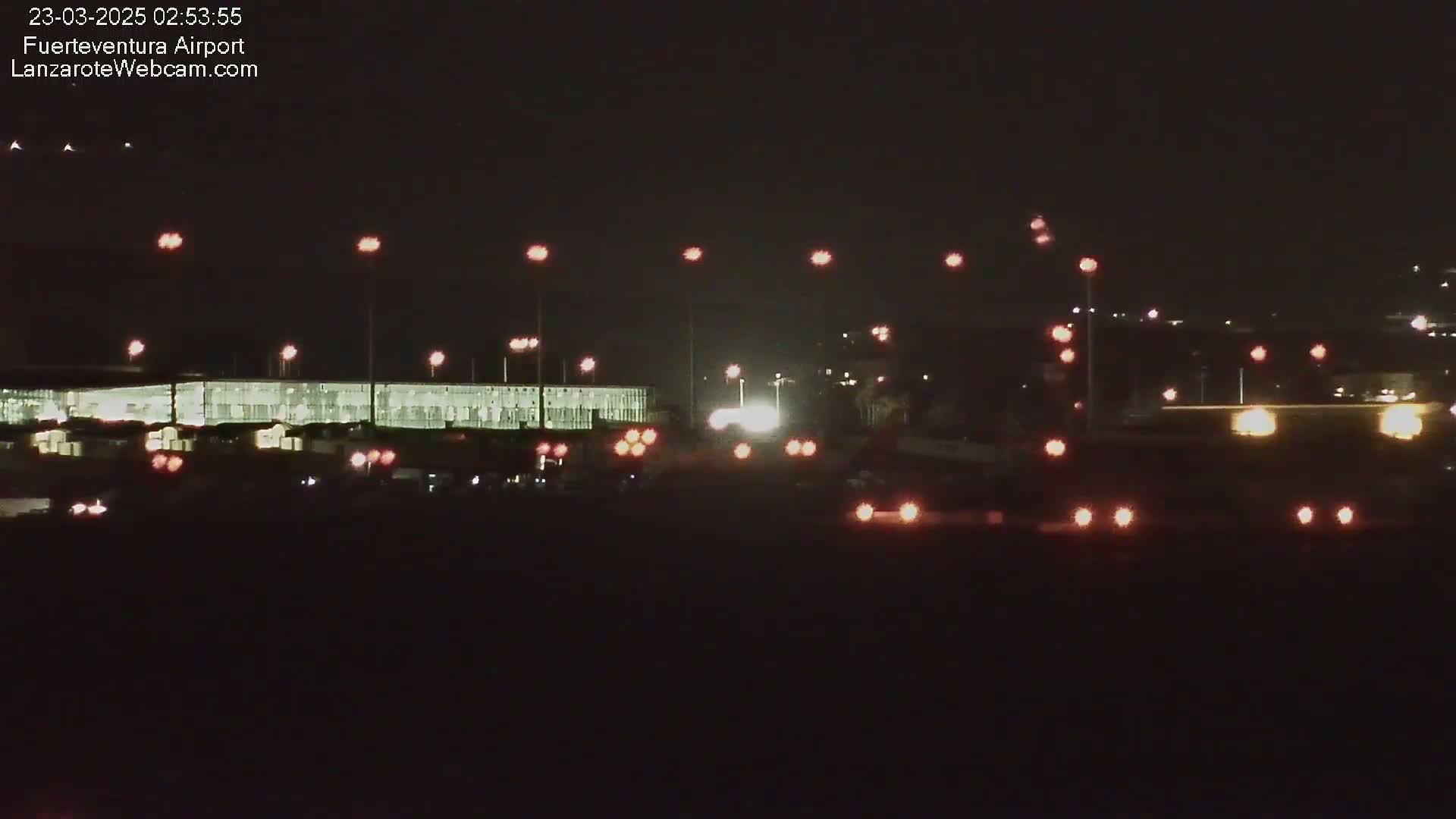 At night, a brightly lit airport terminal building is visible, surrounded by various lights and vehicles on the tarmac.