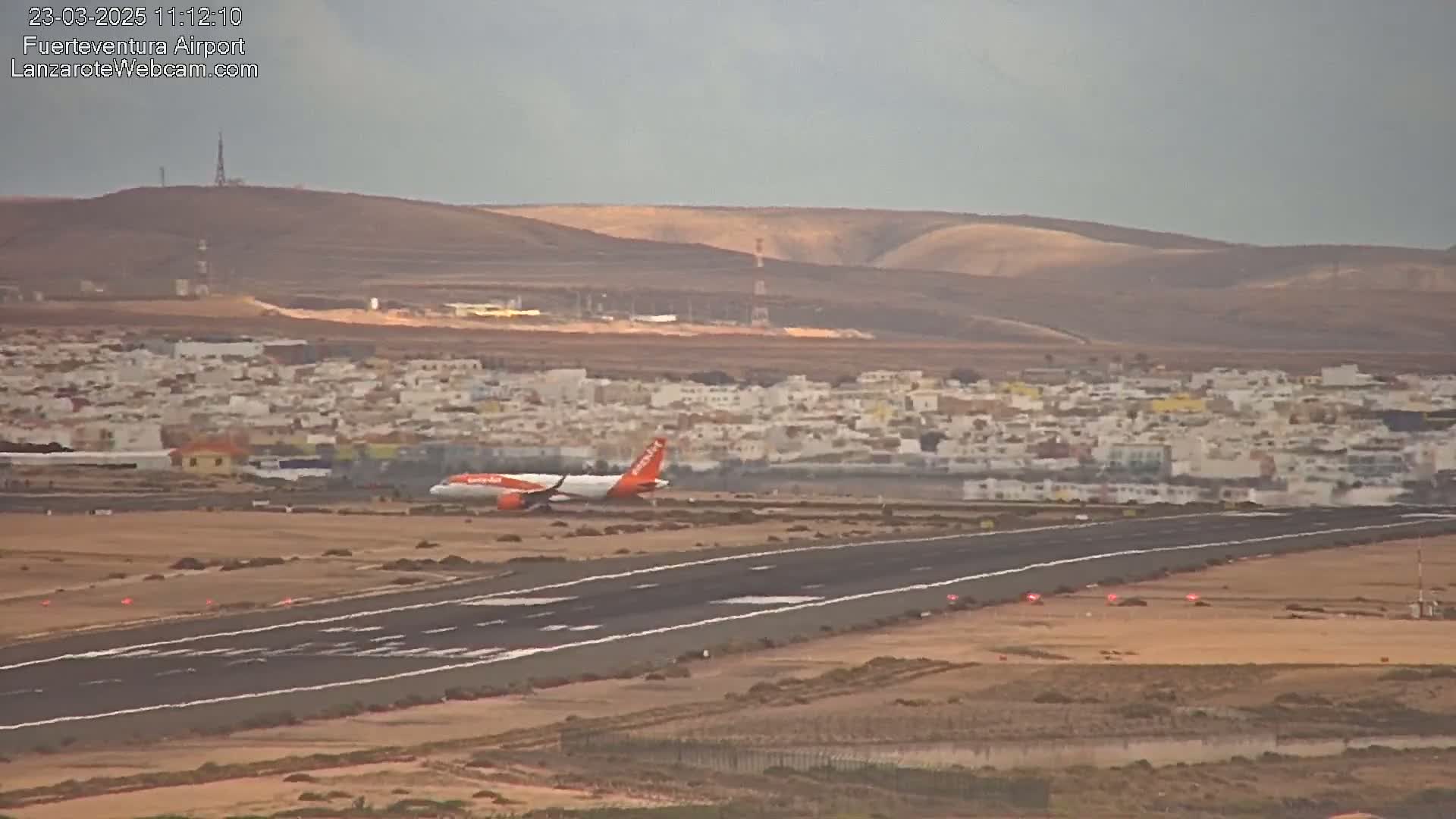 An orange and white airplane taxis on a runway near a town under an overcast sky.