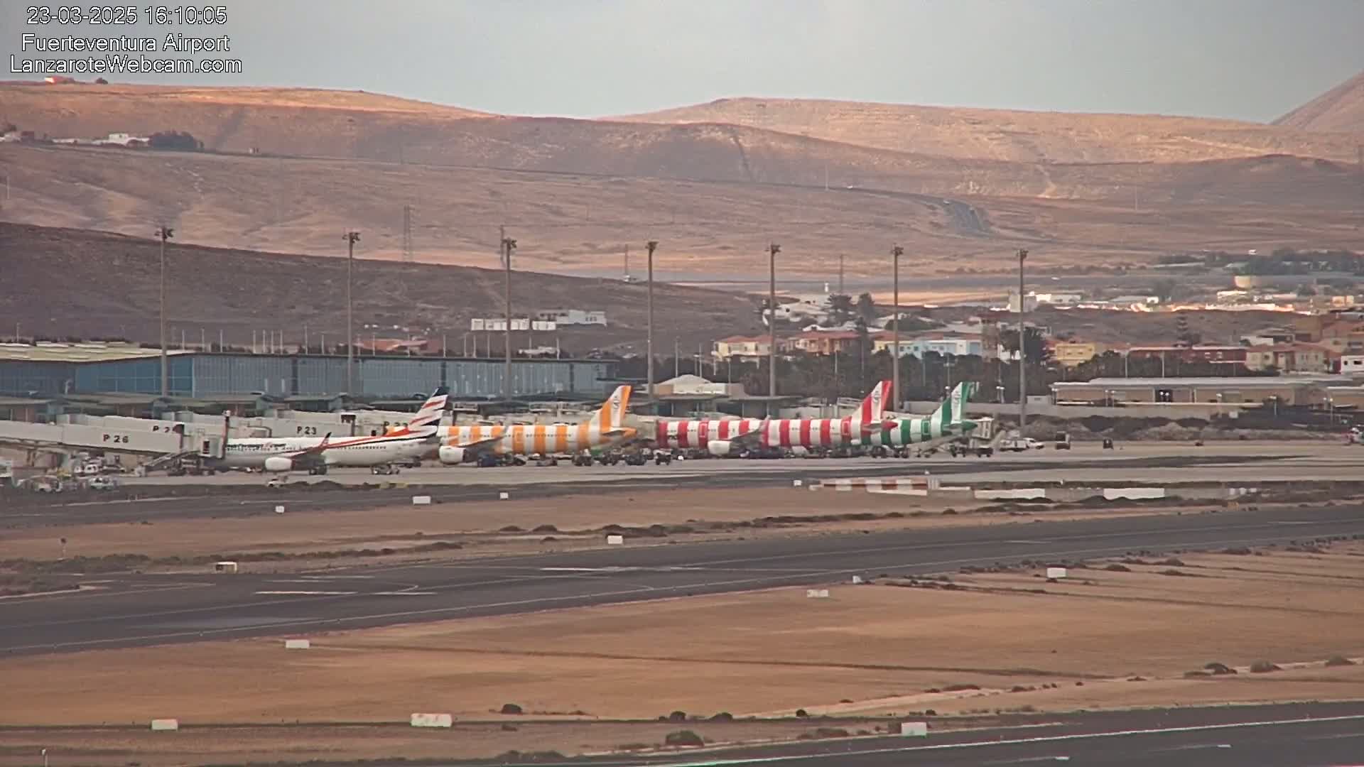Several airplanes are parked at an airport tarmac under an overcast sky, with desert-like terrain visible in the background.