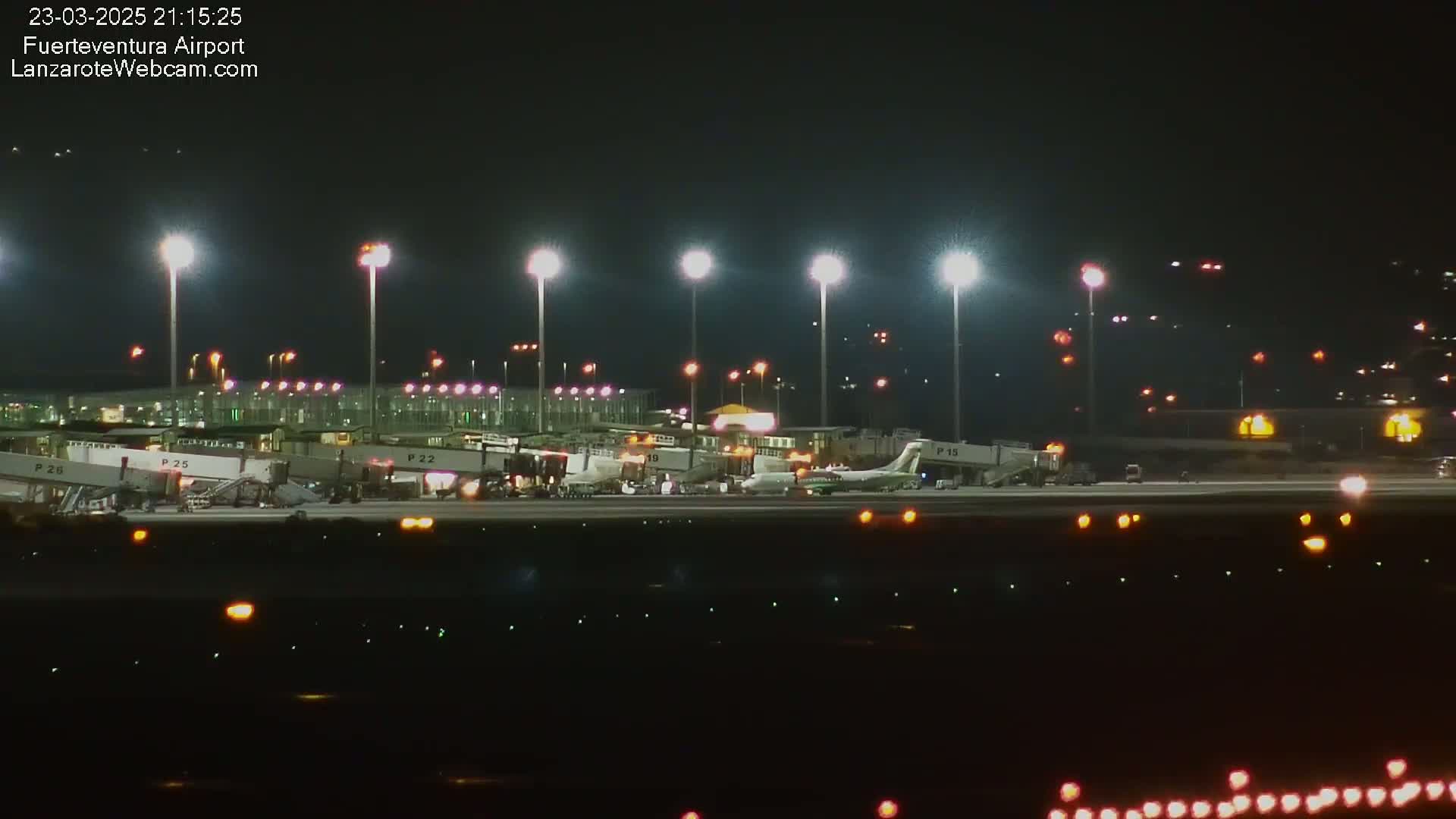 At night, several airplanes are parked at gates at an airport, brightly lit by tall lights, with ground support equipment visible.