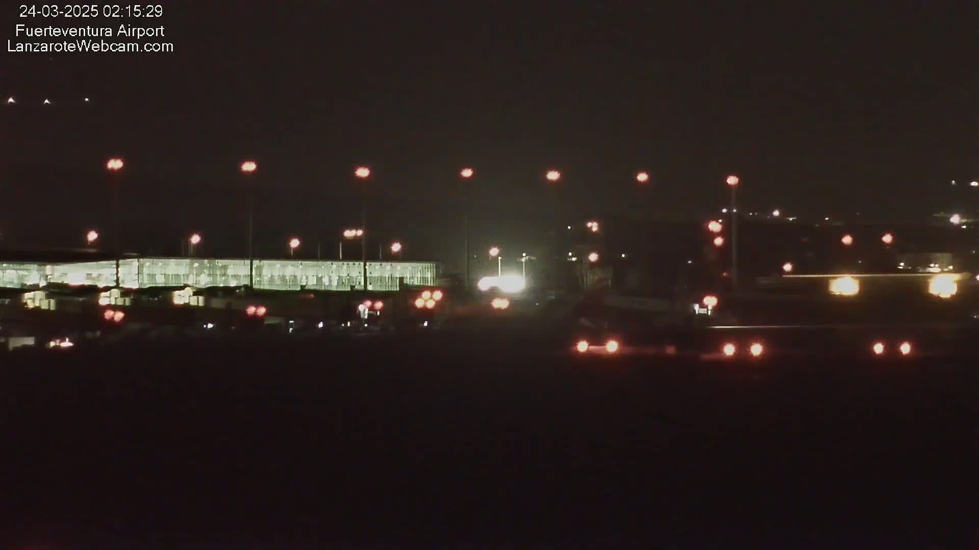 At night, a brightly lit airport terminal building is visible, with various vehicles and lights scattered across the tarmac.