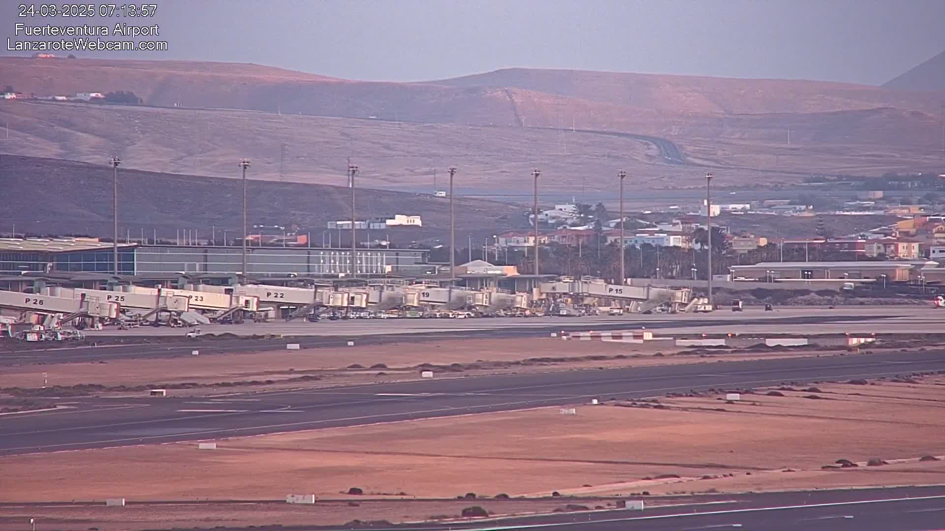An airport tarmac with several jet bridges connected to a terminal building, under a hazy sky and against a backdrop of barren hills.