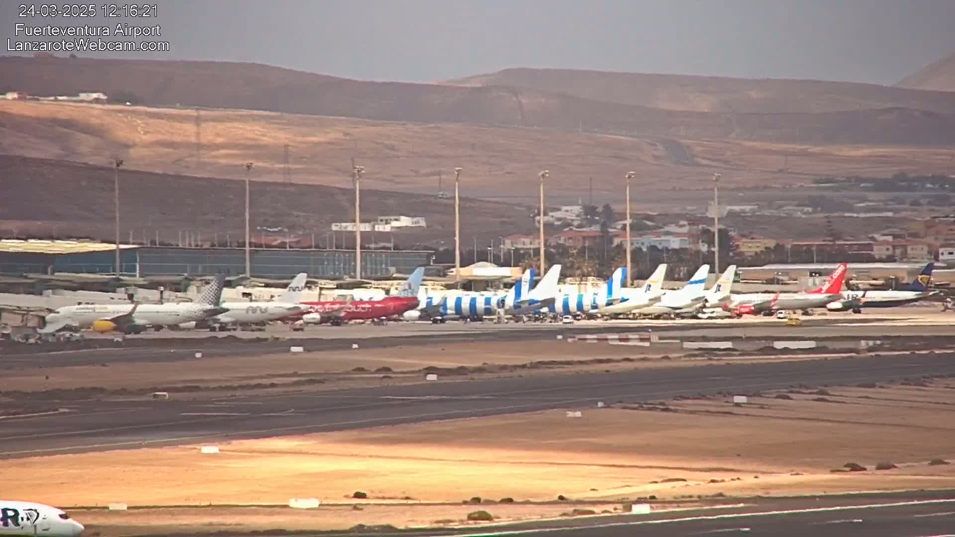 A tarmac with numerous airplanes parked near an airport terminal under a hazy sky, with arid hills visible in the background.