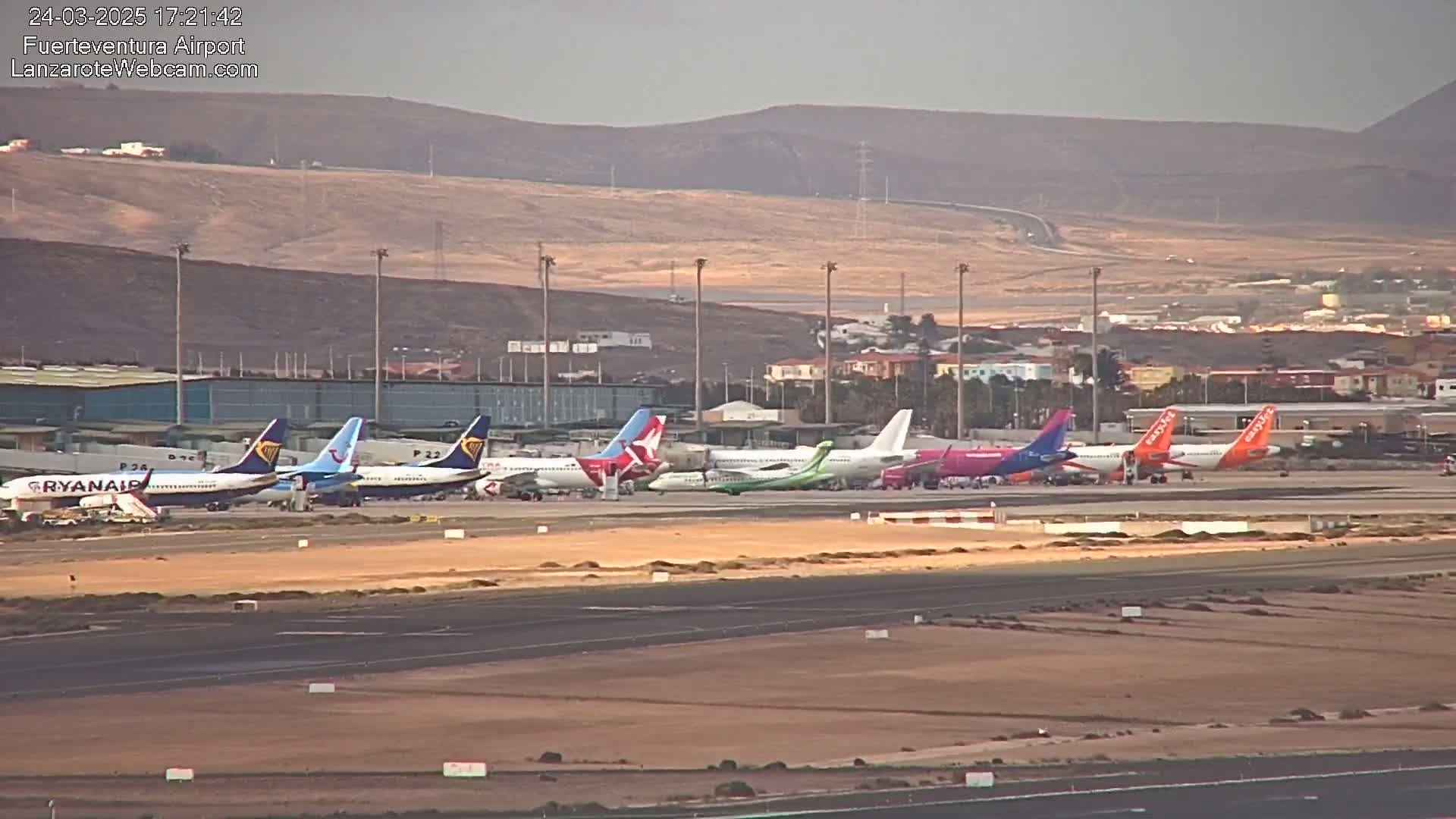 Several airplanes are parked on a tarmac at an airport, under a clear, sunny sky with brown hills in the background.