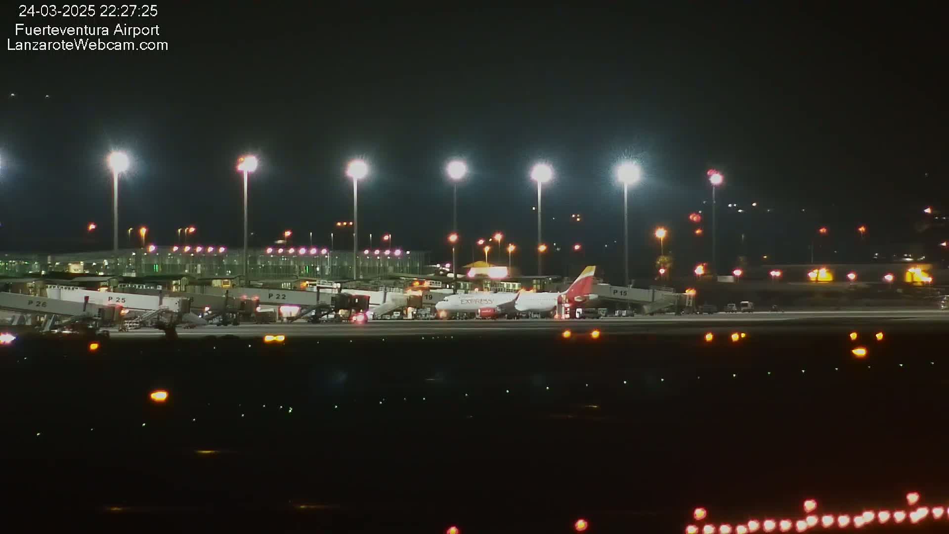 A nighttime view of an airport tarmac with several airplanes parked at gates, illuminated by bright lights.