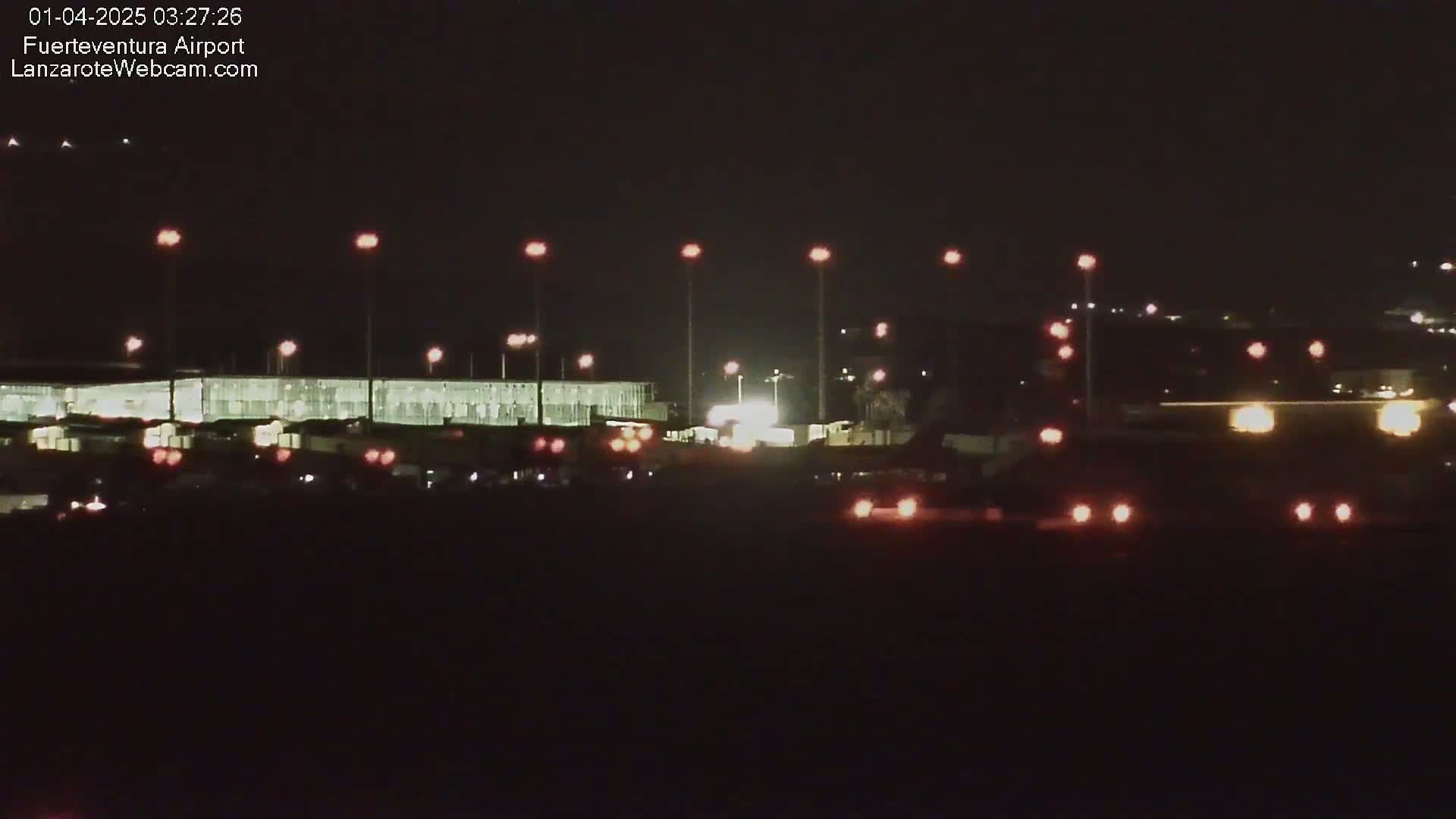 At night, a long, brightly lit building is visible, surrounded by various lights and vehicles, under a dark, clear sky.