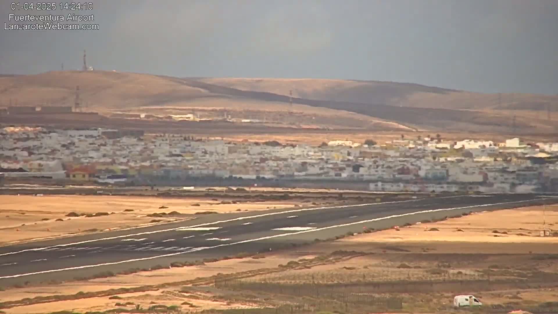A long runway stretches across a flat, sandy landscape toward a distant town nestled against brown hills under a hazy sky.
