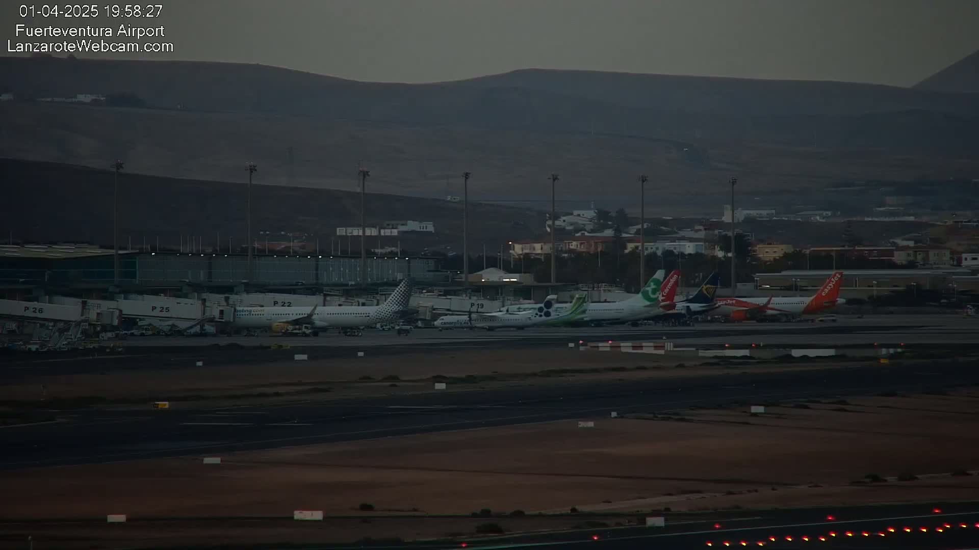 Several airplanes are parked at an airport at dusk, with a dark mountain range in the background.