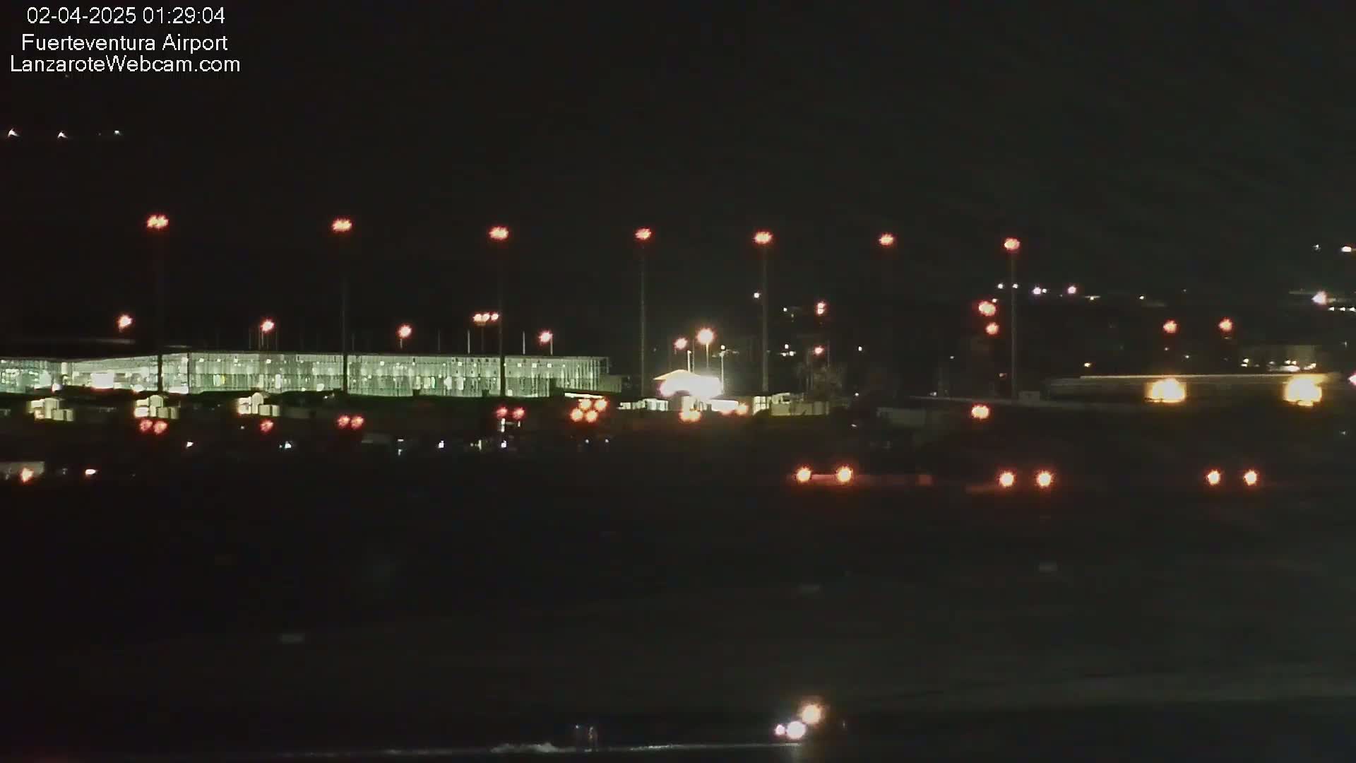 At night, a brightly lit airport terminal is visible, surrounded by numerous lights illuminating the tarmac and surrounding area.
