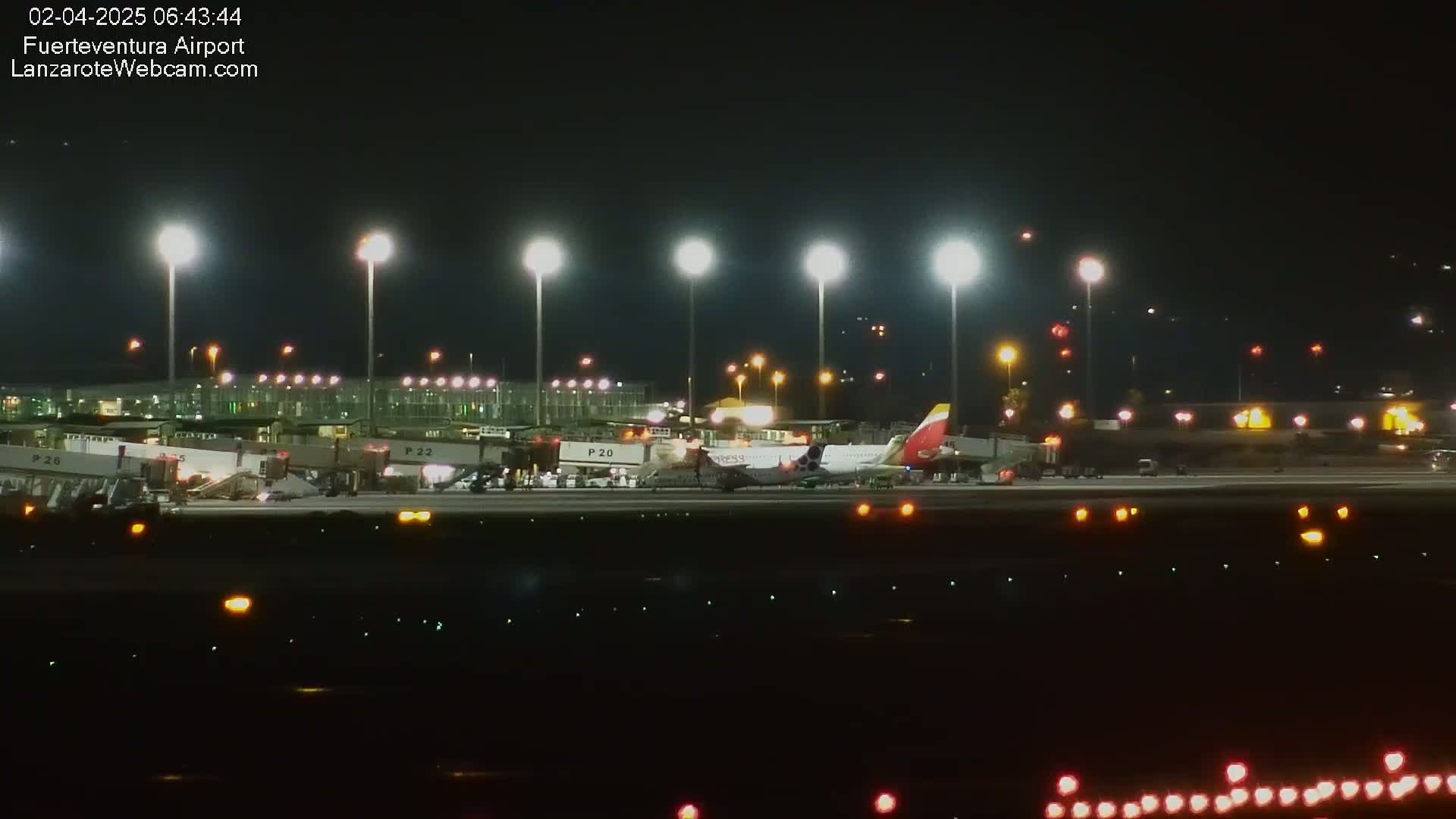 At night, an airplane is parked at a brightly lit airport gate with several jet bridges attached to the terminal.