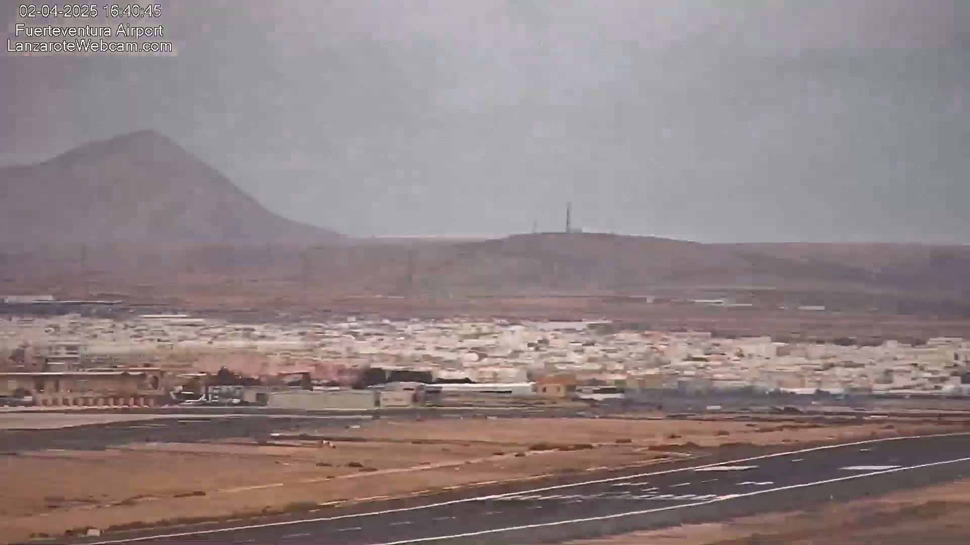 A hazy view shows a town next to an airport runway, with hills and mountains in the background under an overcast sky.