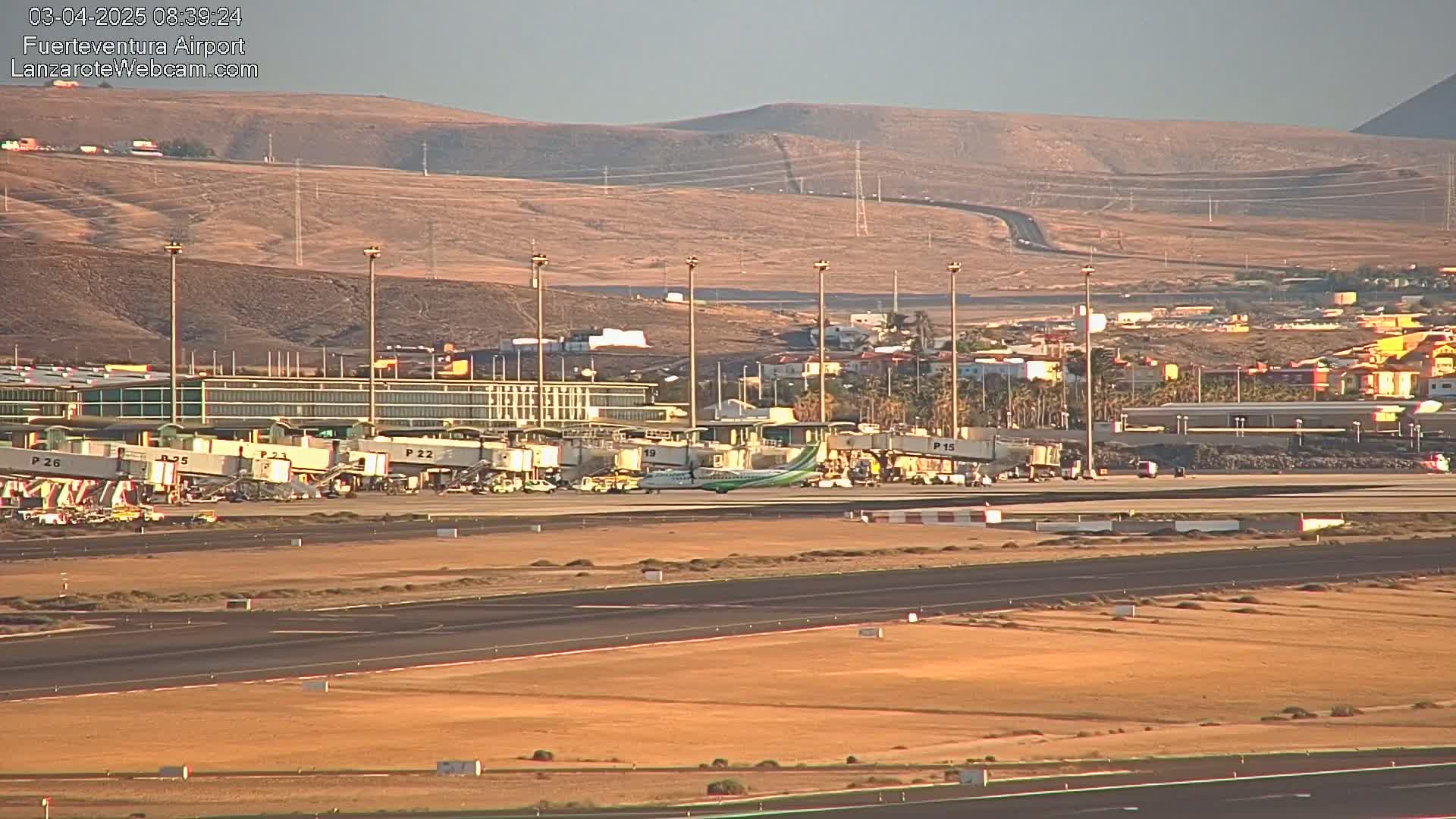 A turboprop airplane taxis on an airport runway under a clear, bright sky near a terminal with several jet bridges.