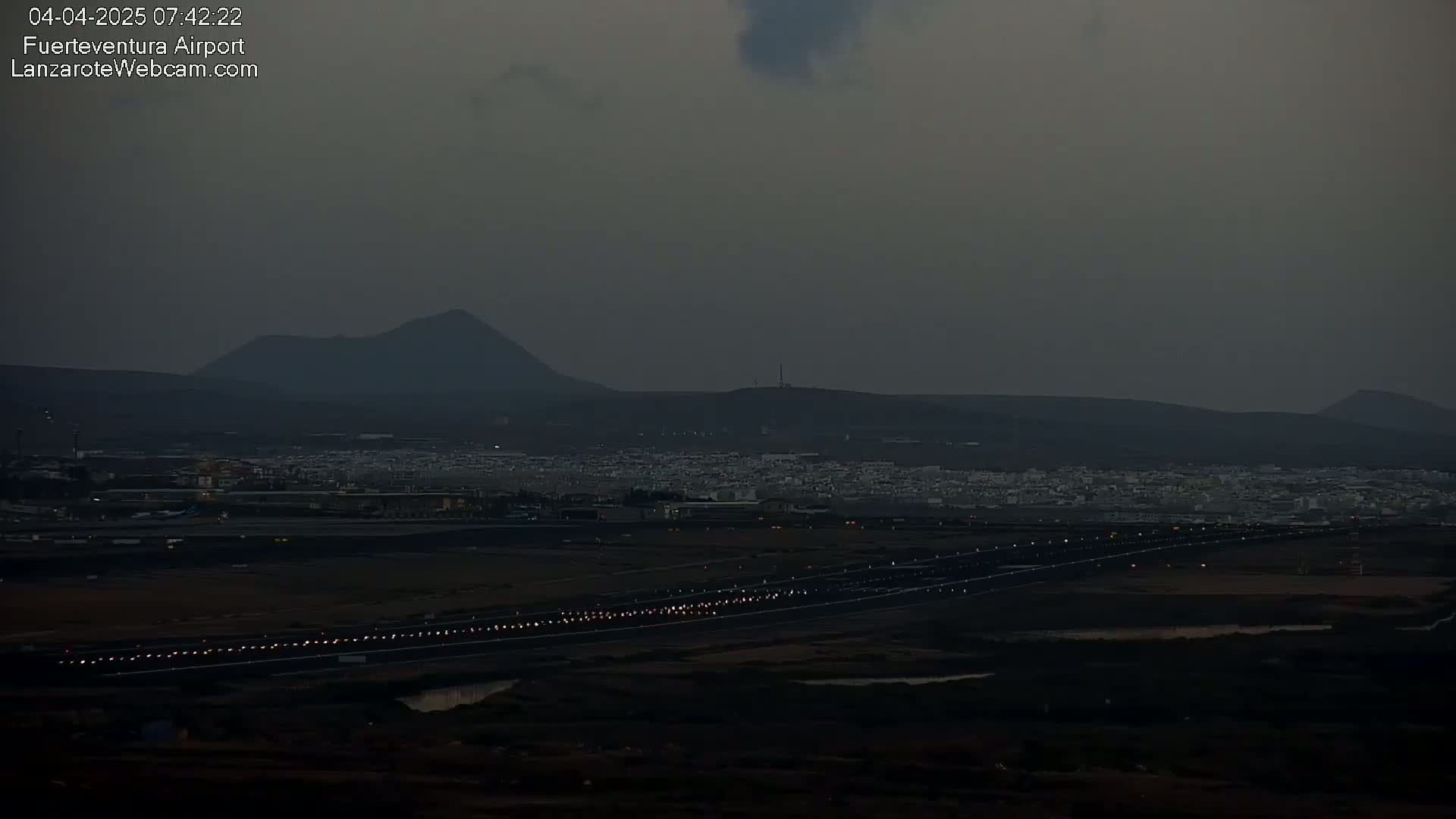 A dark, hazy scene shows an airport runway with lights illuminated, a town nestled below dark volcanic hills in the distance.