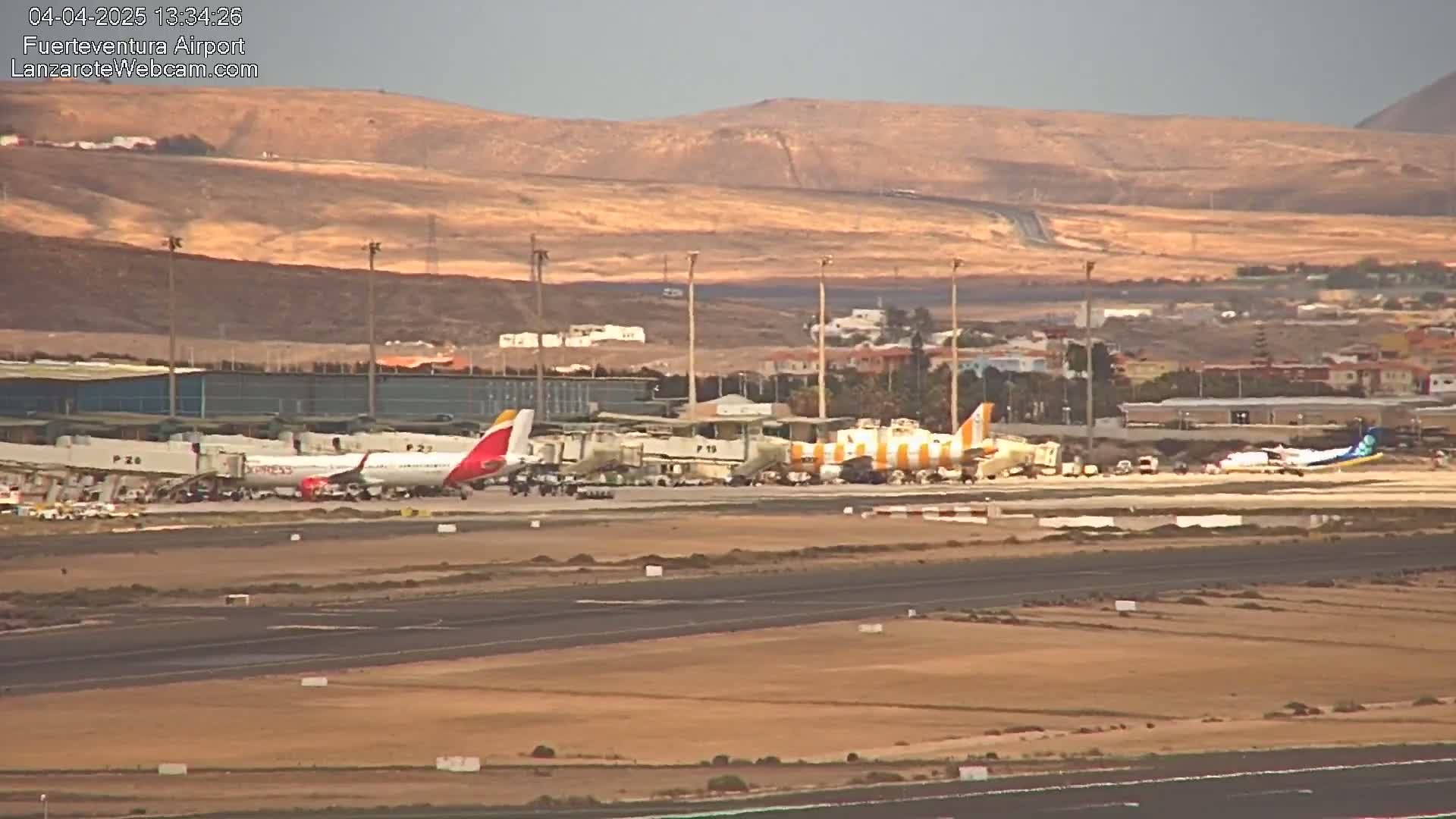 Several airplanes are parked at an airport tarmac in front of an airport terminal building, under a sunny sky with brown hills in the background.