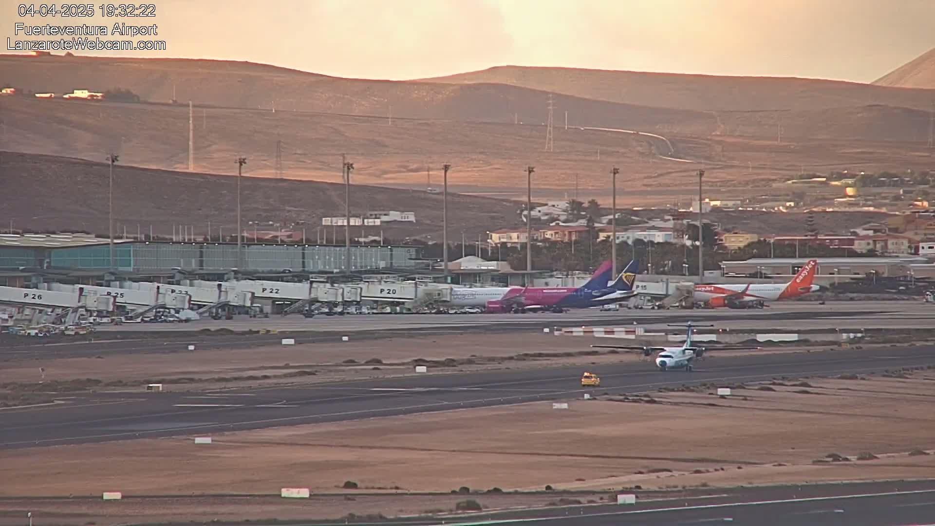 A small propeller plane taxis on a runway at an airport near several larger planes parked at gates under a dusky sky.