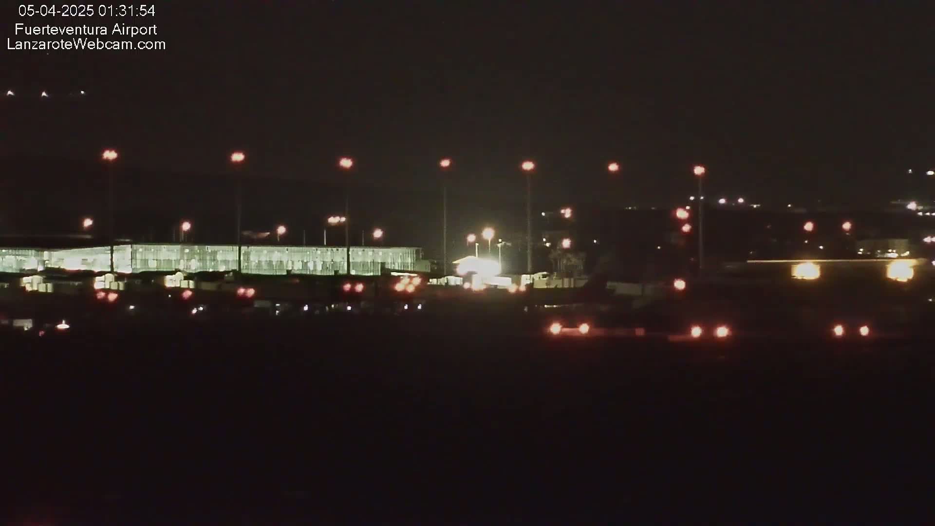 At night, a long, brightly lit airport terminal building is visible, surrounded by numerous lights illuminating the tarmac and surrounding area.