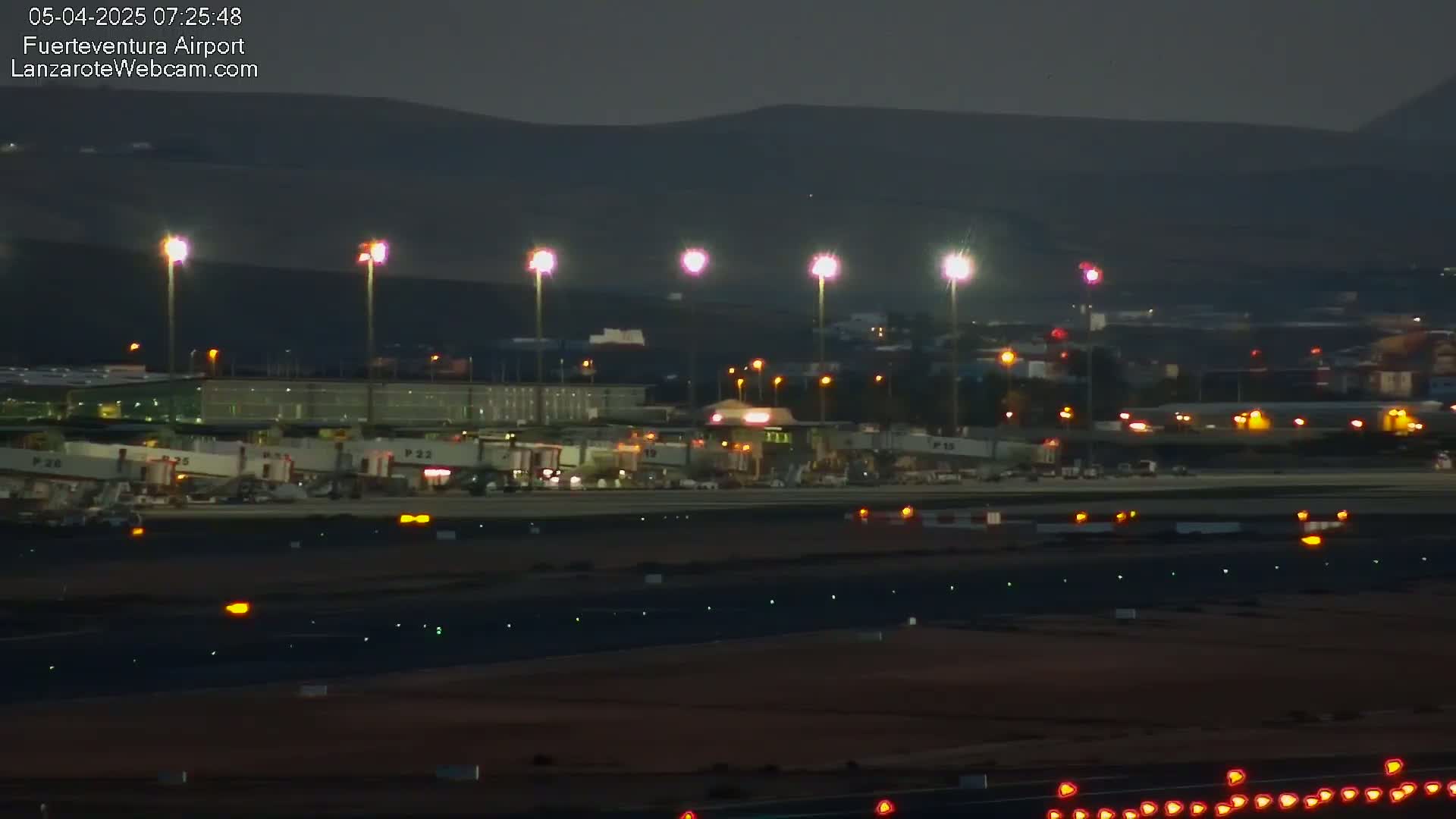 A brightly lit airport terminal at night, with several jet bridges connected to airplanes, is situated in front of dark hills under a clear, dark sky.