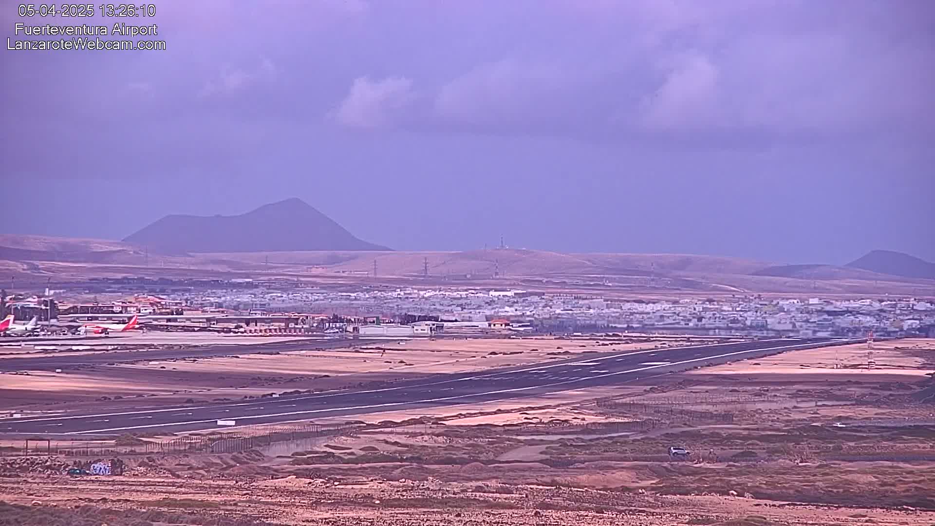 An airport runway stretches across a flat, arid landscape under a mostly cloudy, pale purple sky, with low mountains visible in the distance.