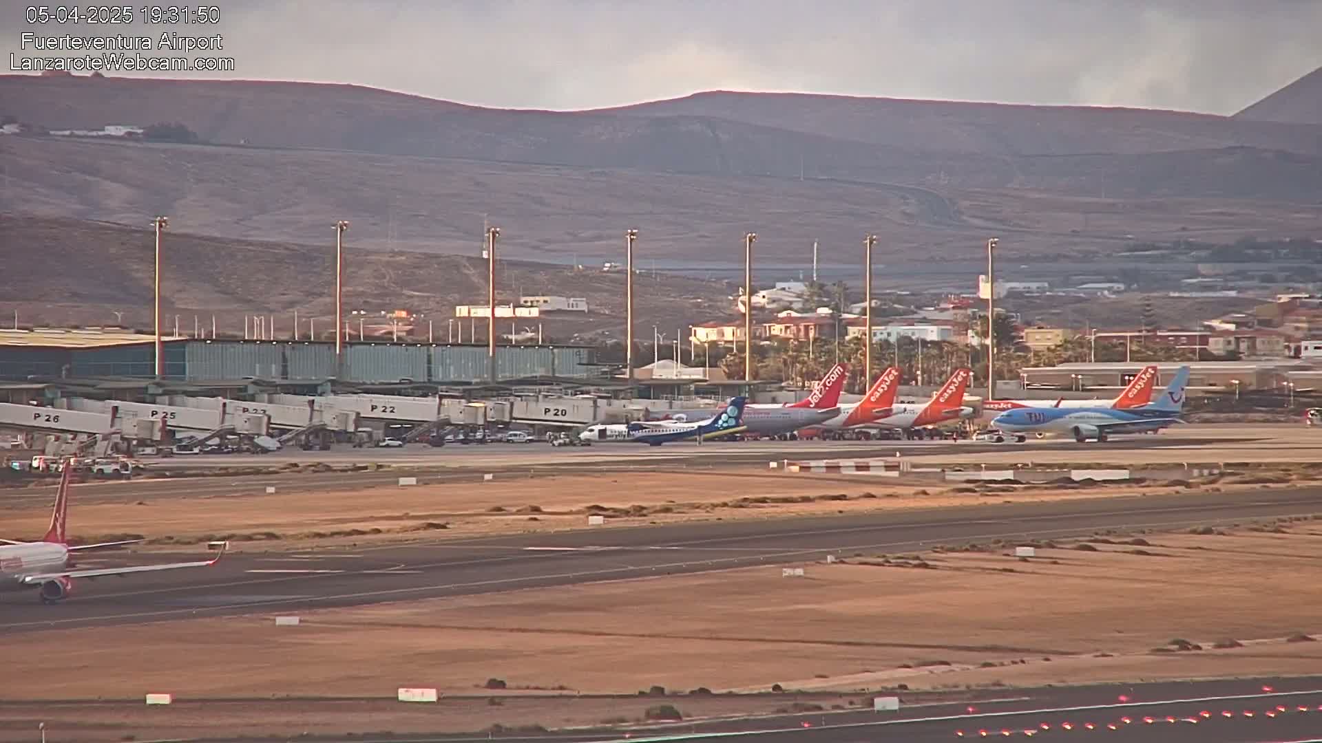 Several airplanes are parked at an airport terminal under a partly cloudy sky, with mountains visible in the background.