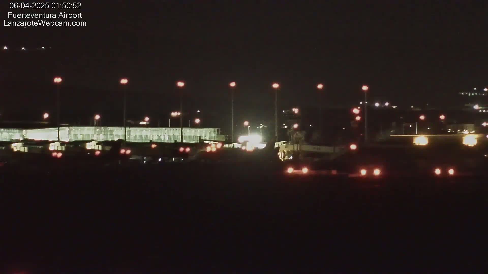 At night, a brightly lit airport terminal is visible, with various vehicles and lights scattered in the foreground and background.