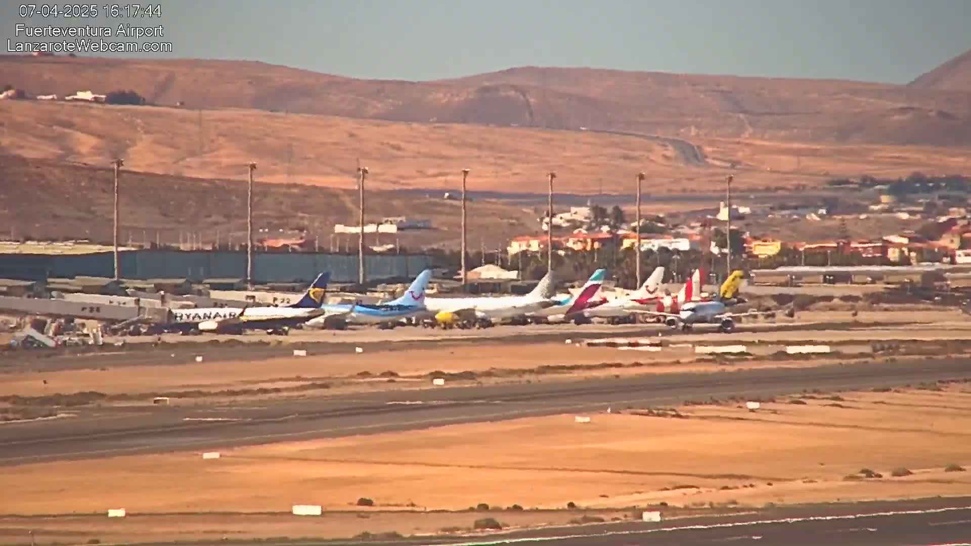 Several airplanes are parked on a tarmac at an airport on a sunny day, with arid, tan-colored hills visible in the background.