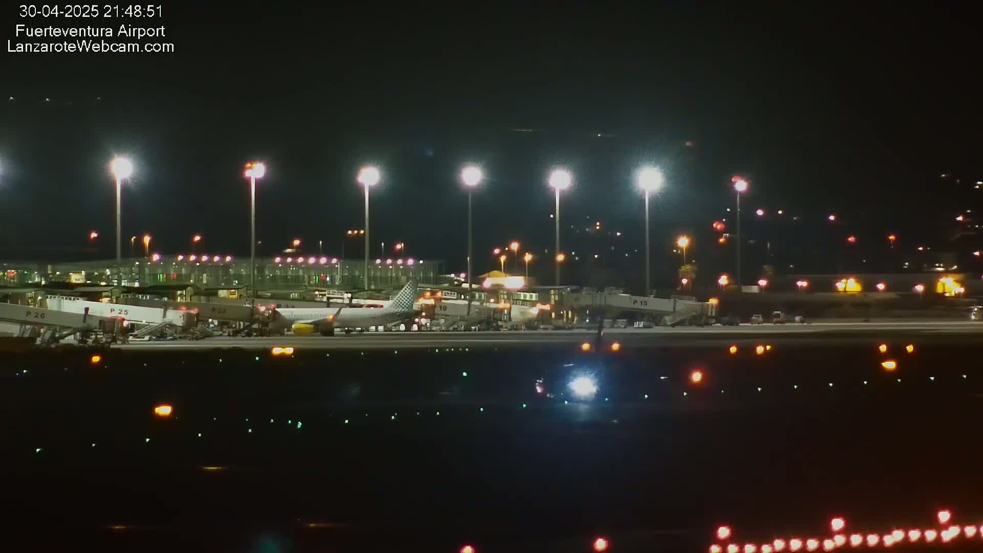 At night, a brightly lit airport tarmac shows several airplanes parked at gates with ground support equipment, and lights illuminating the runway.