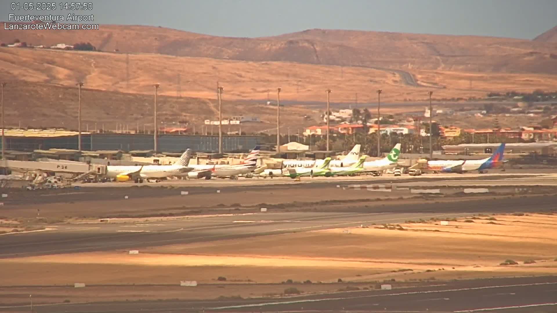 Several airplanes are parked at an airport tarmac near a hilly, desert-like landscape under a mostly clear sky.