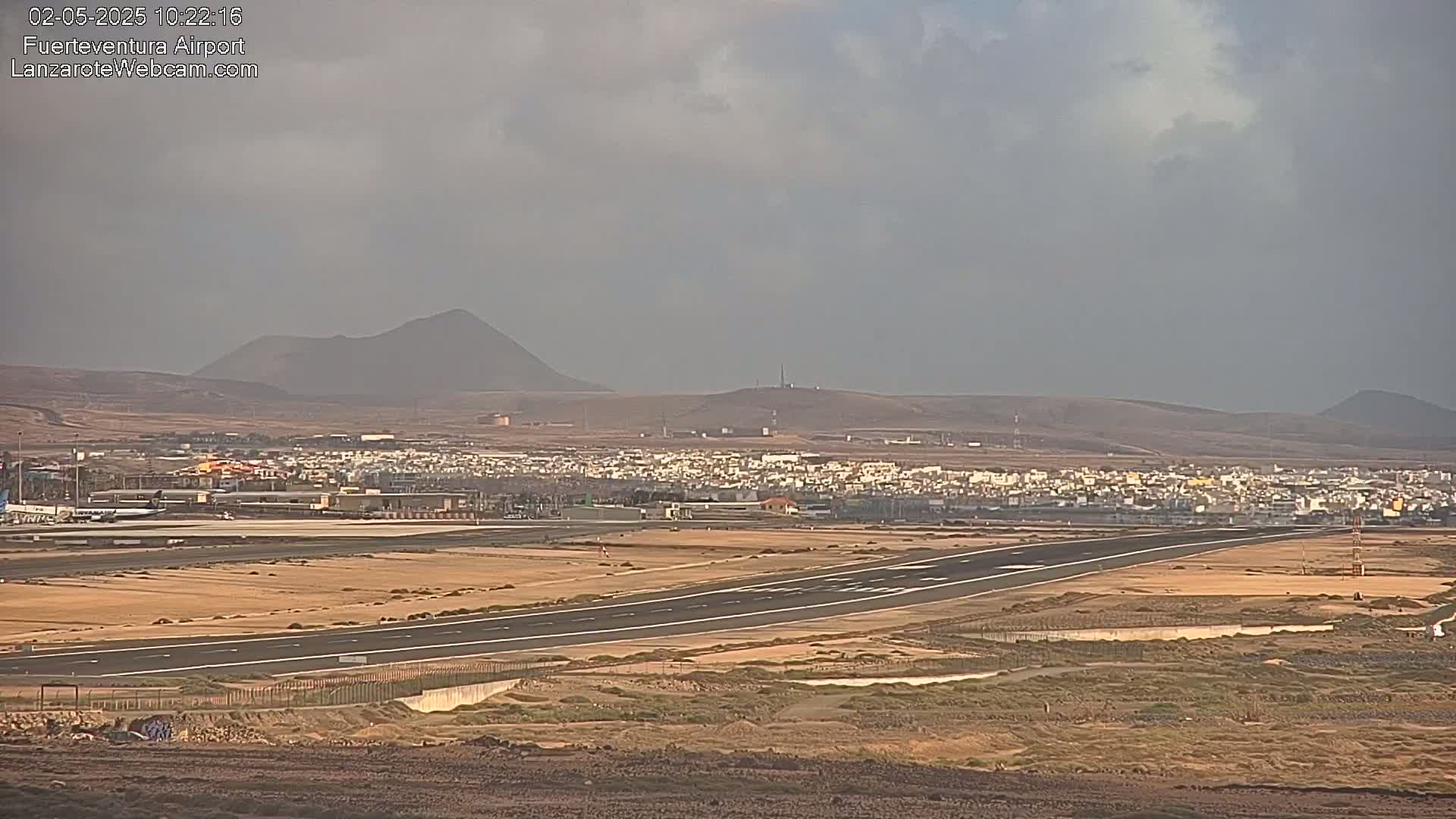 A long runway at an airport stretches across a flat, arid landscape under an overcast sky, with low, volcanic hills in the background.
