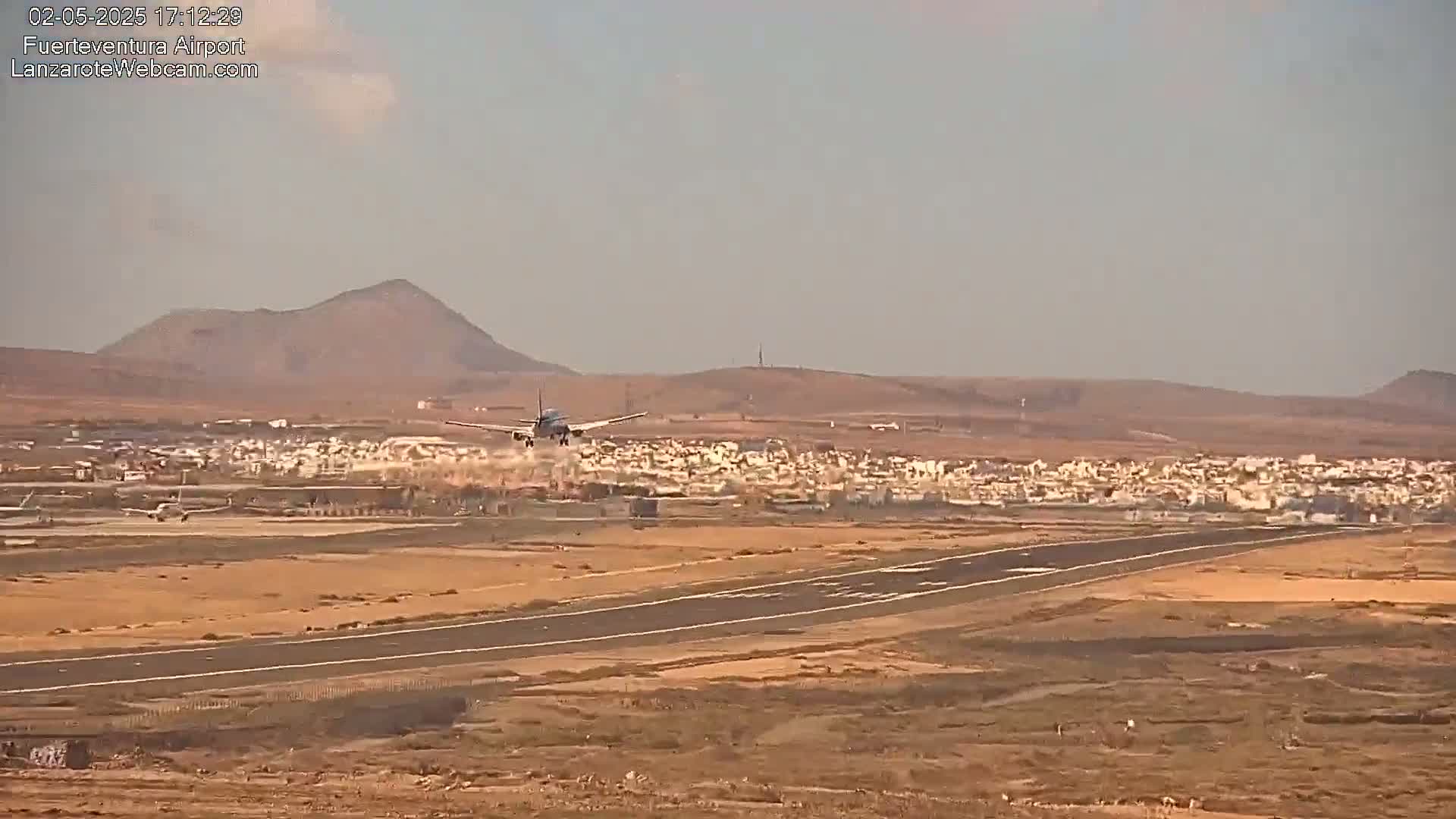 A passenger plane is landing on a runway near a town and low hills under hazy, sunny conditions.