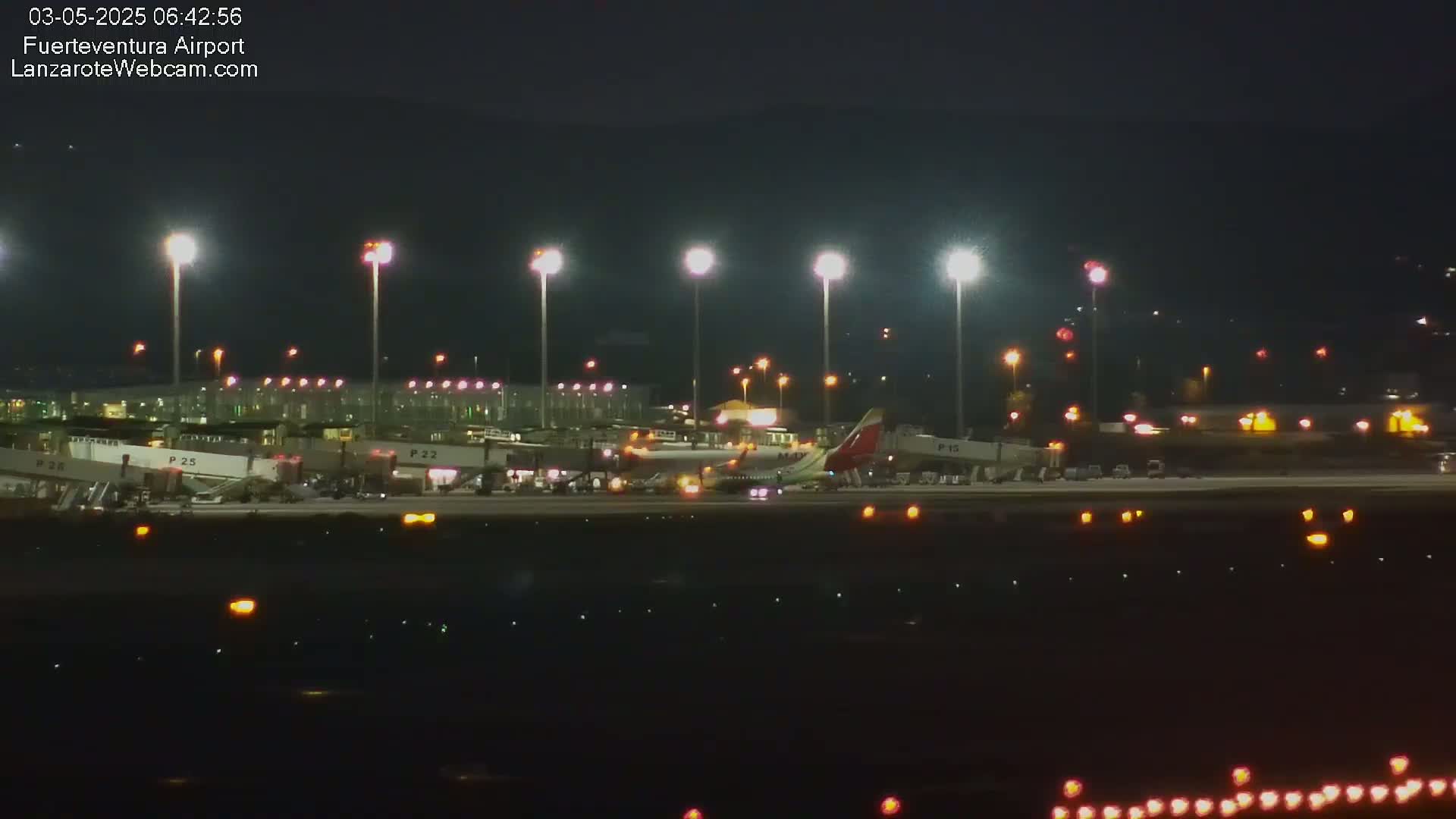 A brightly lit airport at night shows several airplanes parked at gates, with ground support equipment visible.