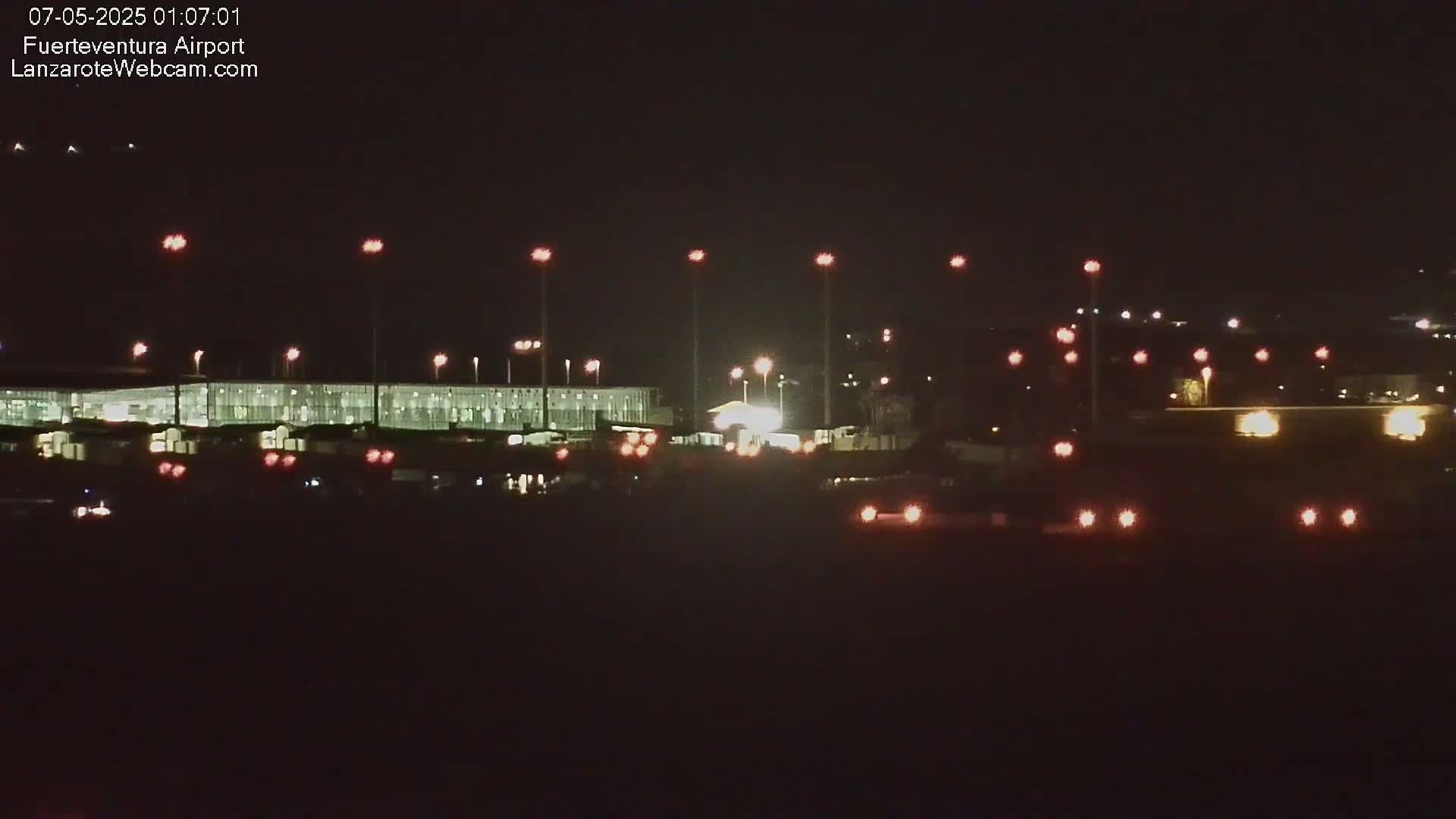 At night, a brightly lit airport terminal is visible, surrounded by numerous lights on tall poles and ground-level lights.