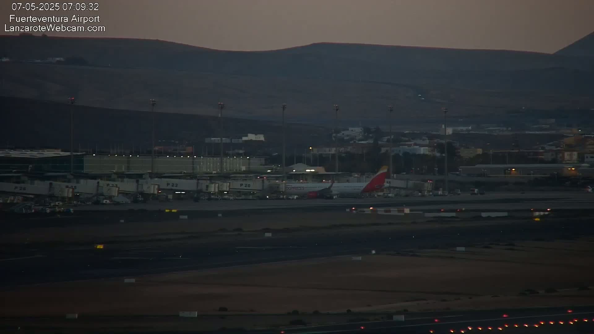A passenger airplane sits at a gate at an airport at dusk, with dark hills visible in the background.