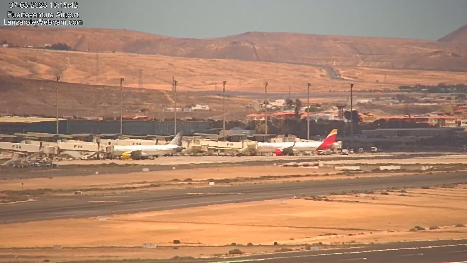 Several airplanes are parked at gates at an airport on a sunny day, with a hilly, desert-like landscape in the background.