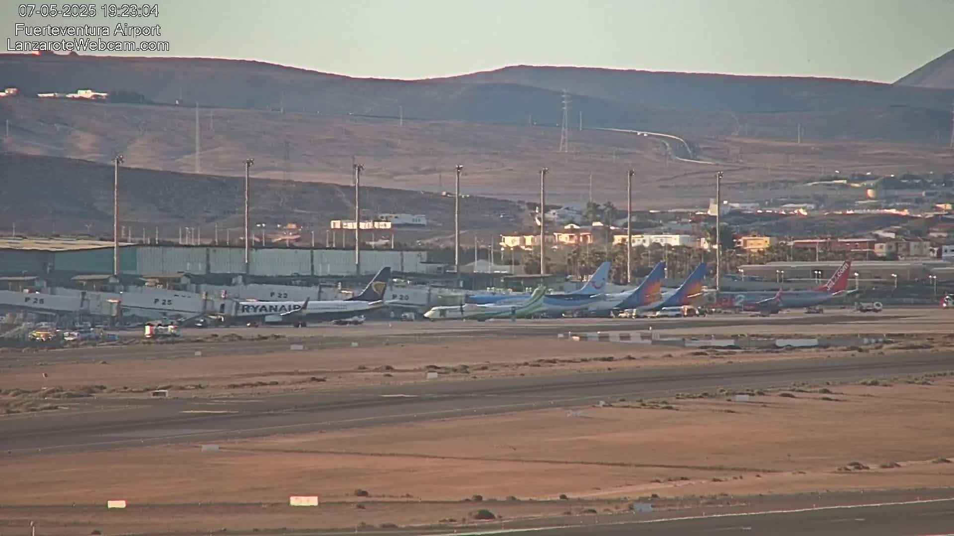 Several airplanes are parked at an airport terminal against a backdrop of dry, hilly terrain under a clear sky.