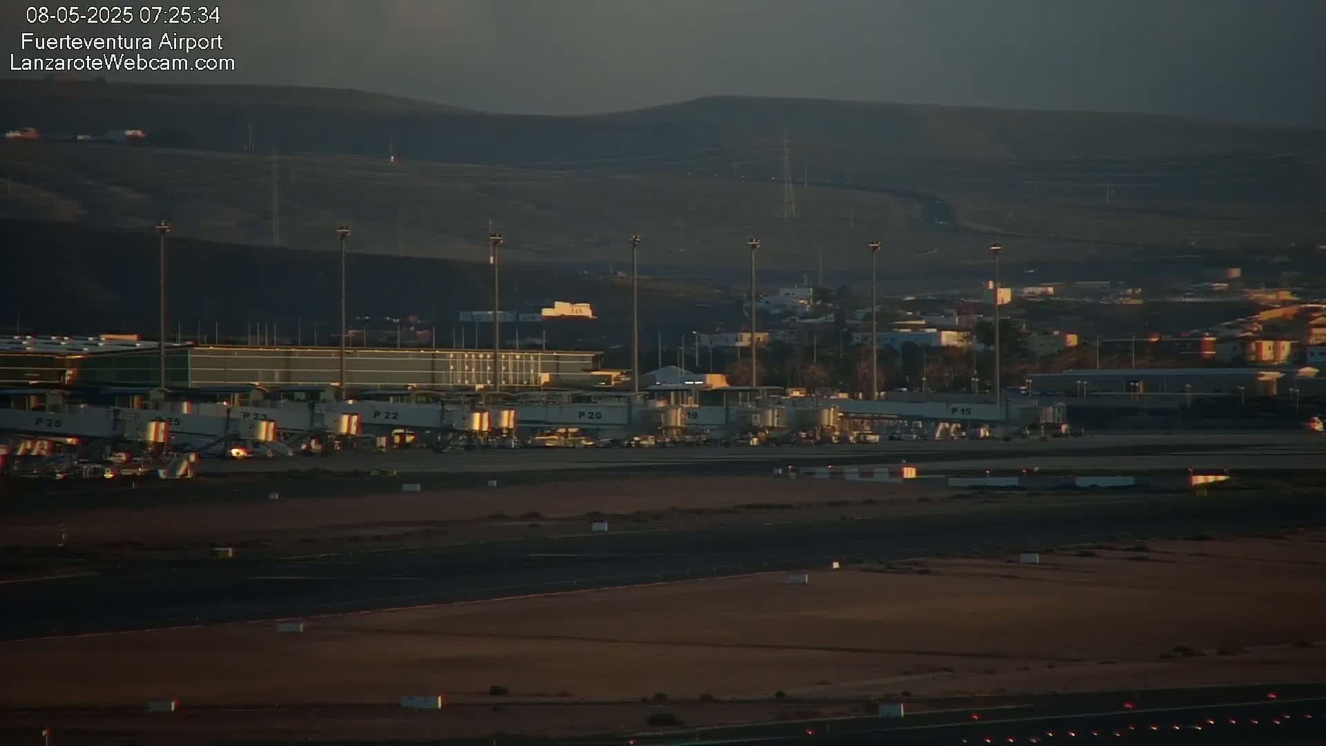 An airport terminal with several jet bridges is visible under a dusky sky, with hills in the background.